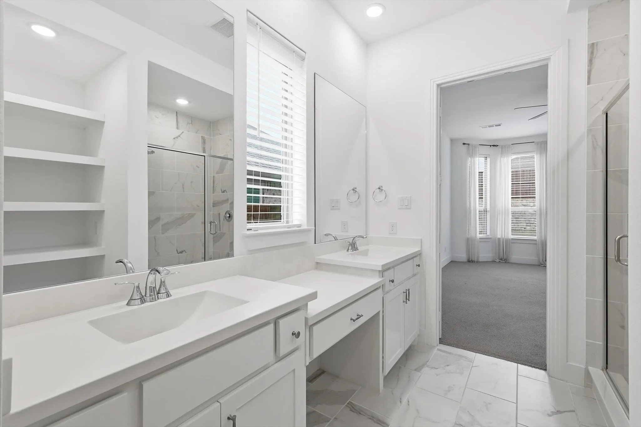 Sleek white cabinetry with marble ceramic tile, oversized walk in shower, dual sinks and make up vanity with natural light and open concept shelving for decor or storage