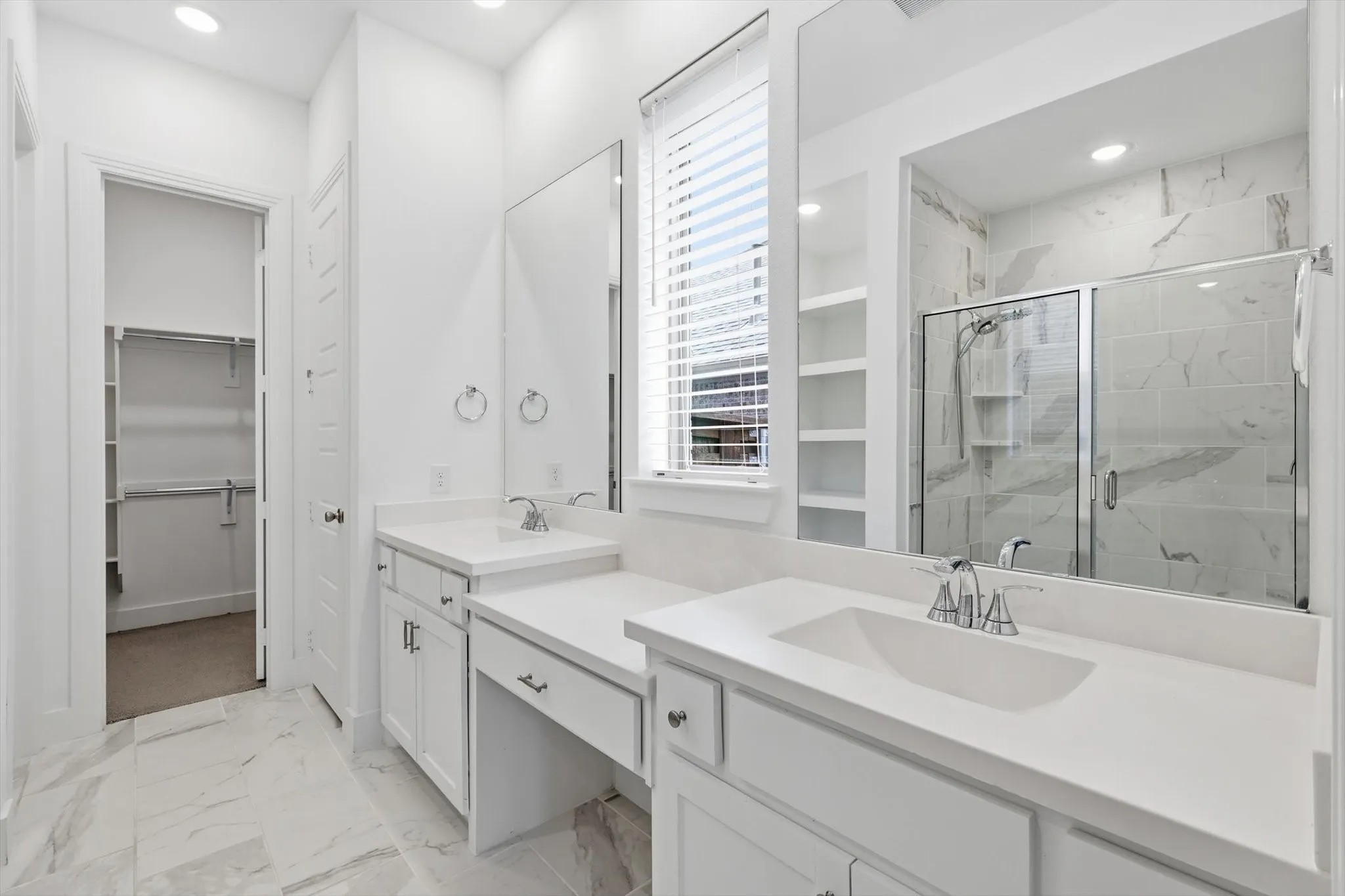 Sleek white cabinetry with marble ceramic tile, oversized walk in shower, dual sinks and make up vanity with natural light and open concept shelving for decor or storage