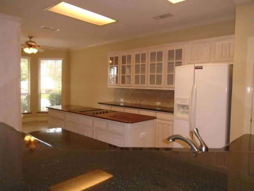 Kitchen featuring glass insert cabinets, white refrigerator with ice dispenser, dark stone counters, tasteful backsplash, and a ceiling fan