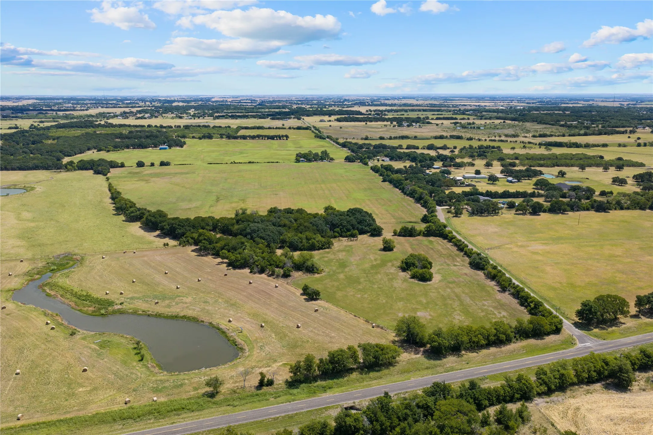 Expansive agricultural land featuring a natural pond, scattered hay bales, mature trees, and a paved road frontage
