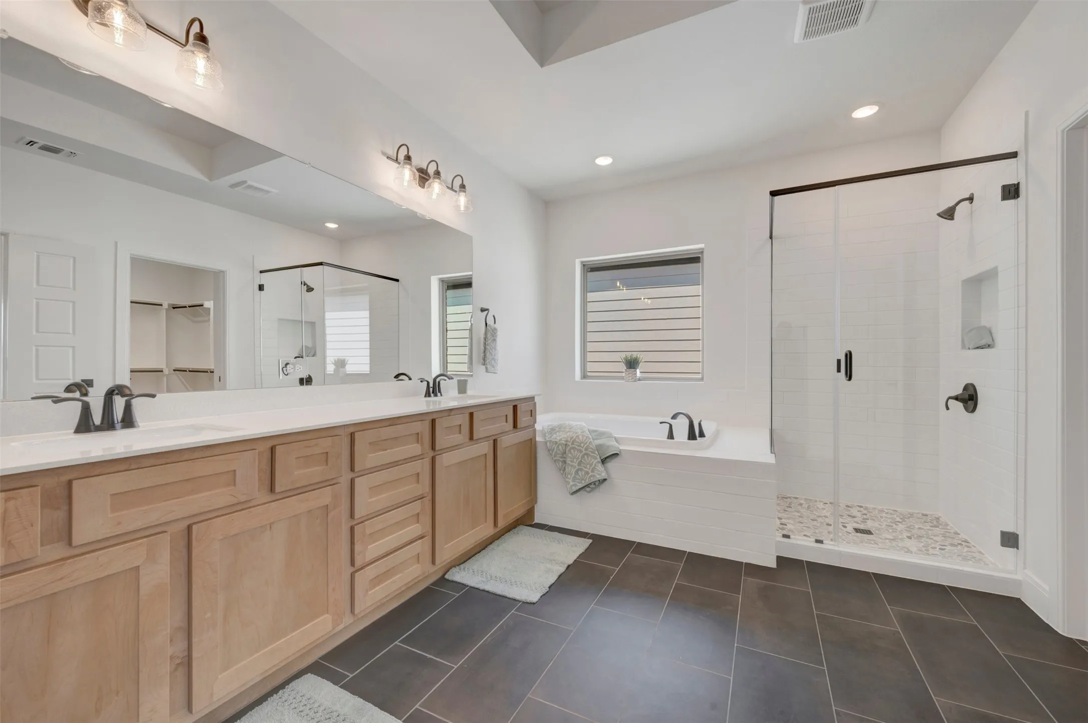 Dual vanity with light wood-finish cabinetry, built-in soaking tub, glass-enclosed shower with pebble-tile floor, large format tile flooring, and wall-mounted light fixtures