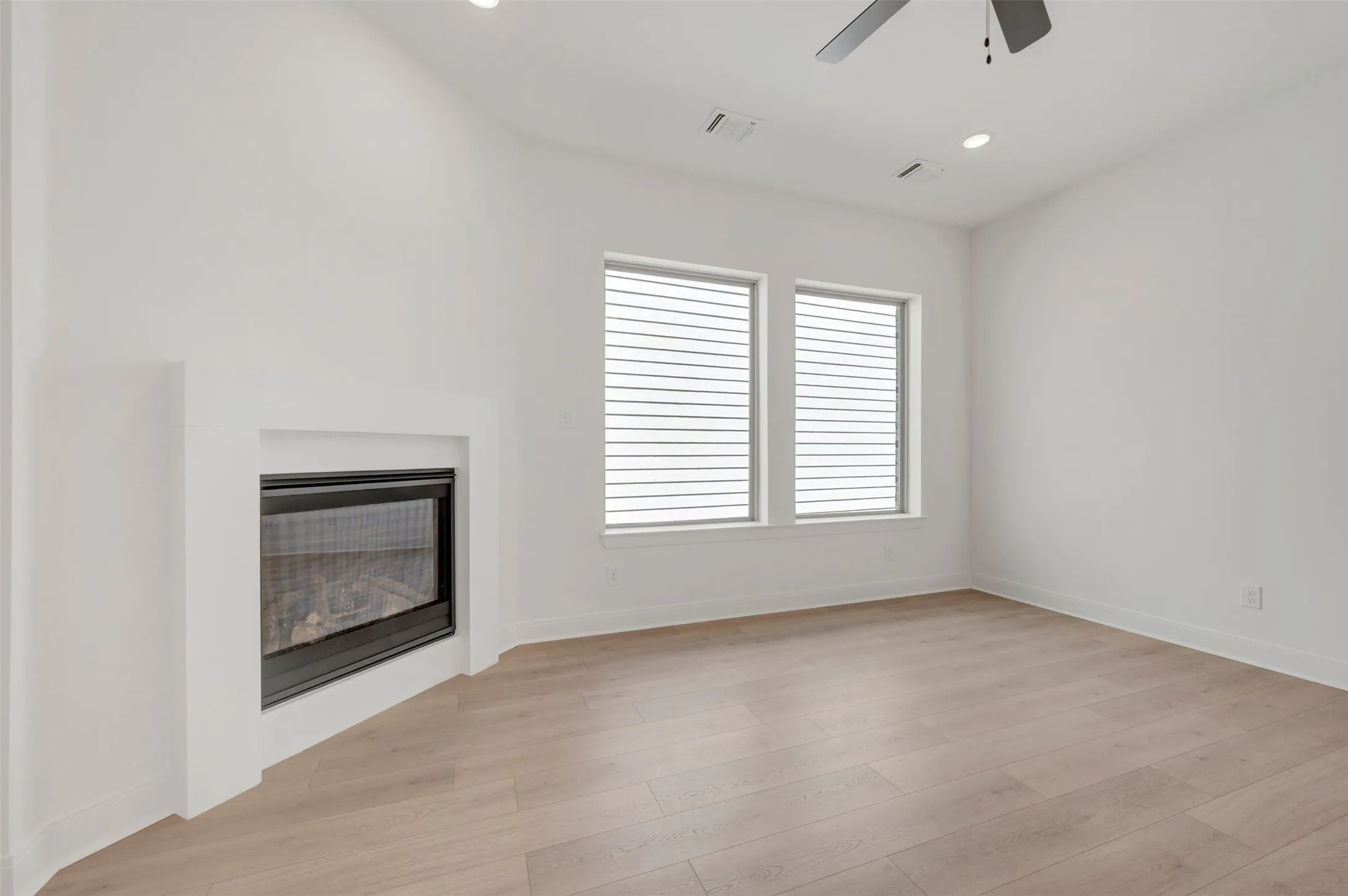 Integrated fireplace with a clean surround, two windows with blinds, wood-finish flooring, and a ceiling fan