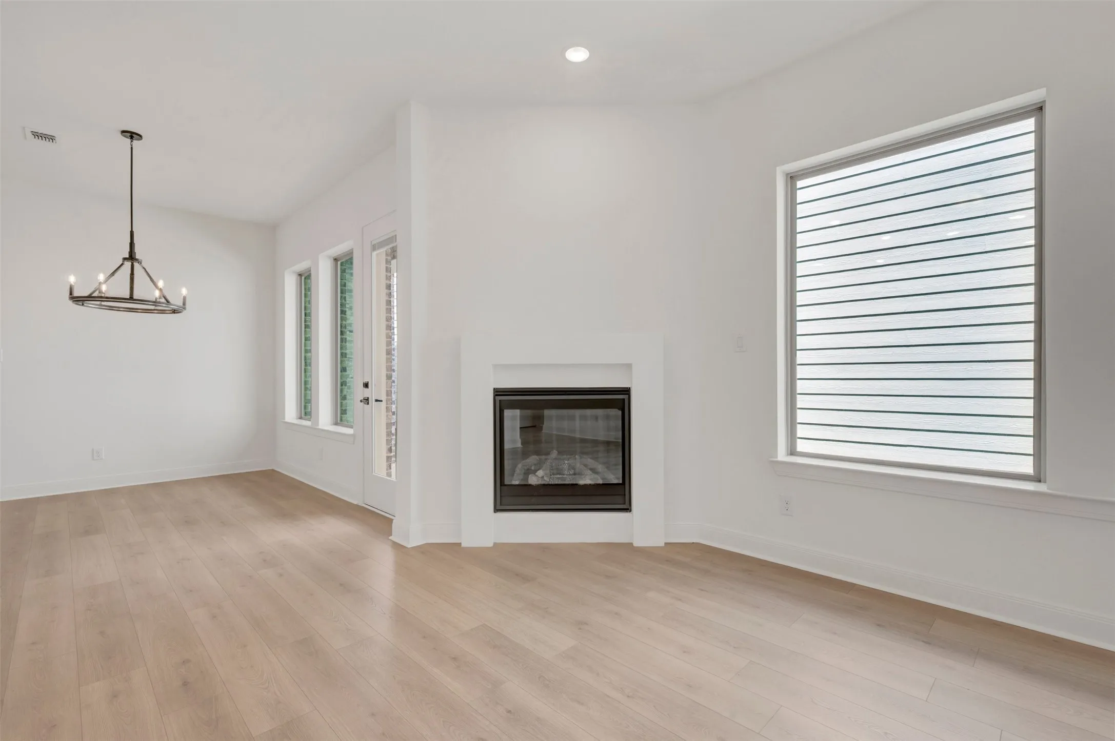 Light wood-finish flooring, corner fireplace with white mantel, large window with horizontal blinds, recessed lighting, and a modern chandelier