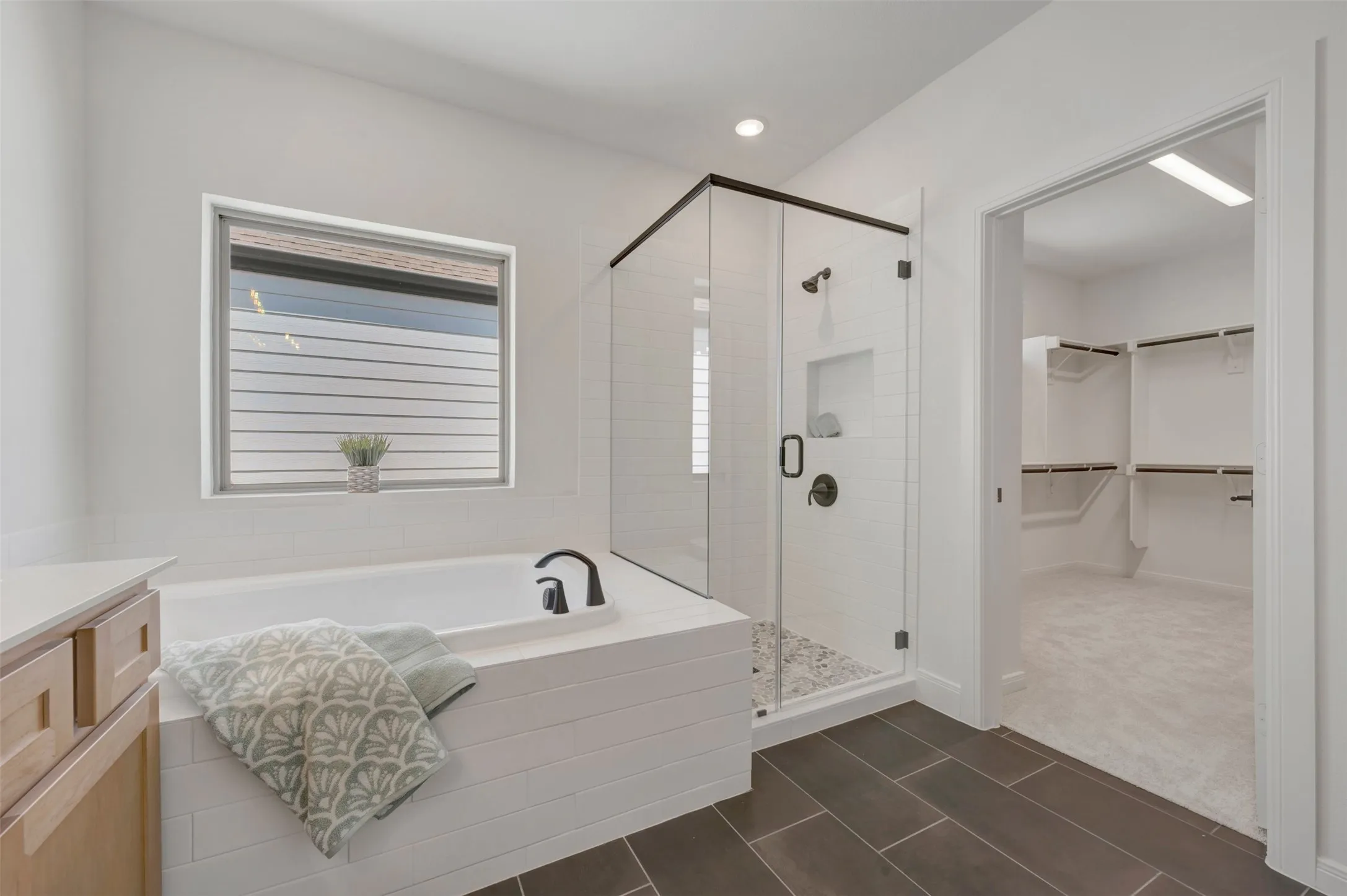 Spa-inspired primary bathroom featuring a freestanding soaking tub with integrated deck space, a frameless glass-enclosed walk-in shower with rainfall showerhead and tile surround, dual vanity with wood-finish cabinetry, and slate-look tile flooring throughout
