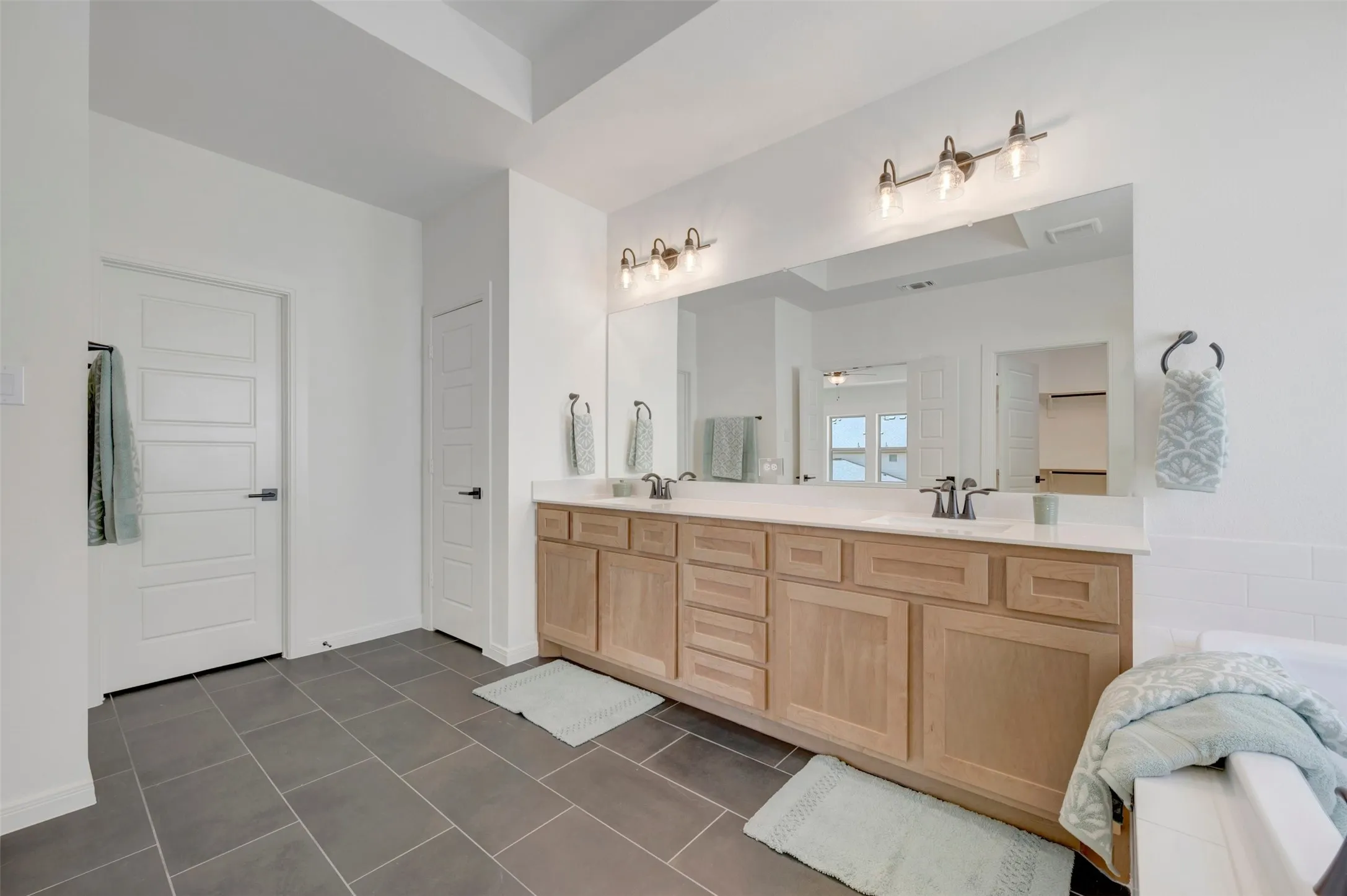 Dual vanity with light wood-finish cabinetry and white countertop, large wall-mounted mirror, brushed nickel light fixtures, and dark tile flooring