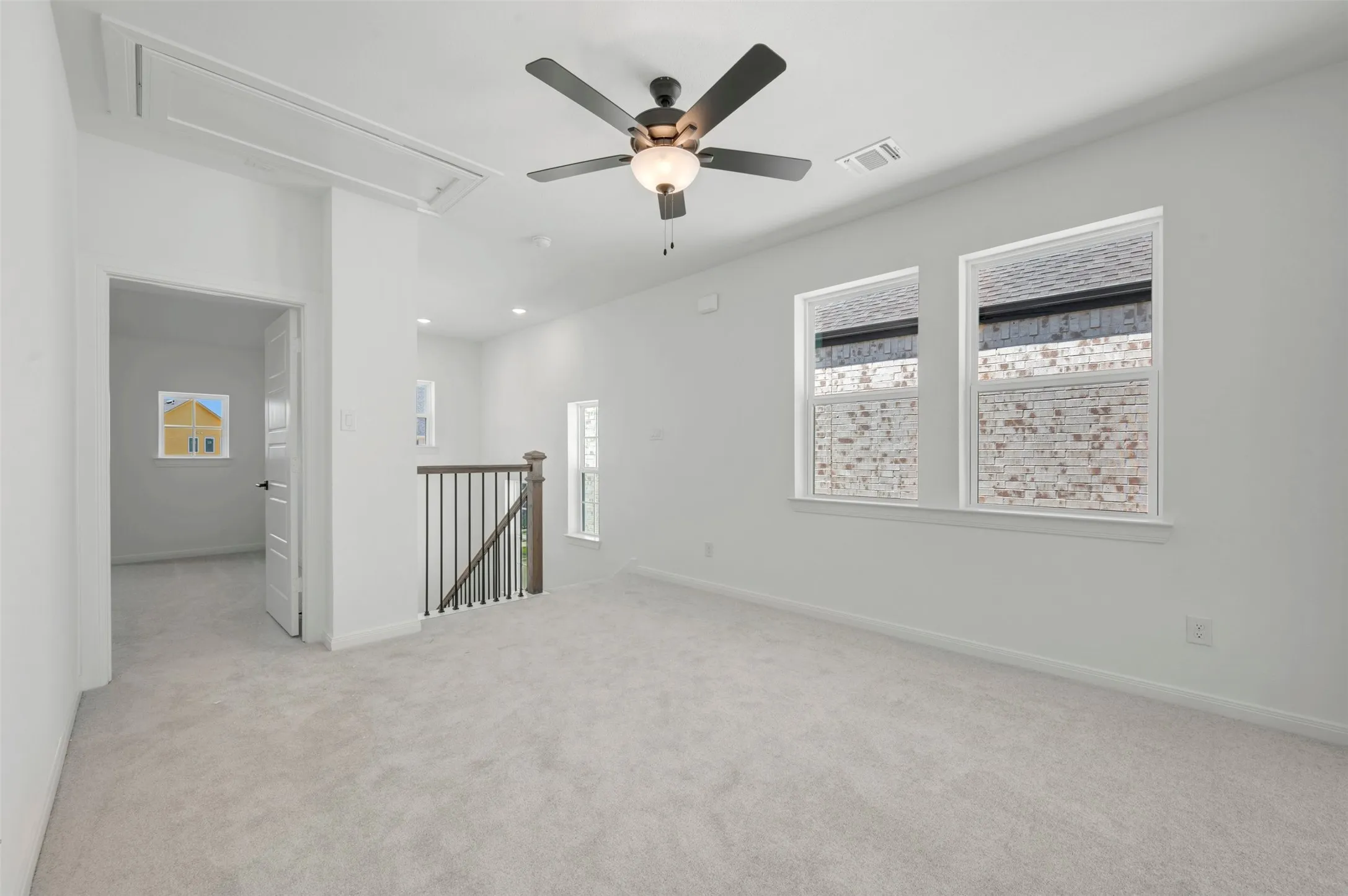 Bright, open loft space featuring neutral carpet flooring, ceiling fan with integrated lighting, expansive windows with cellular shades, and a railed staircase