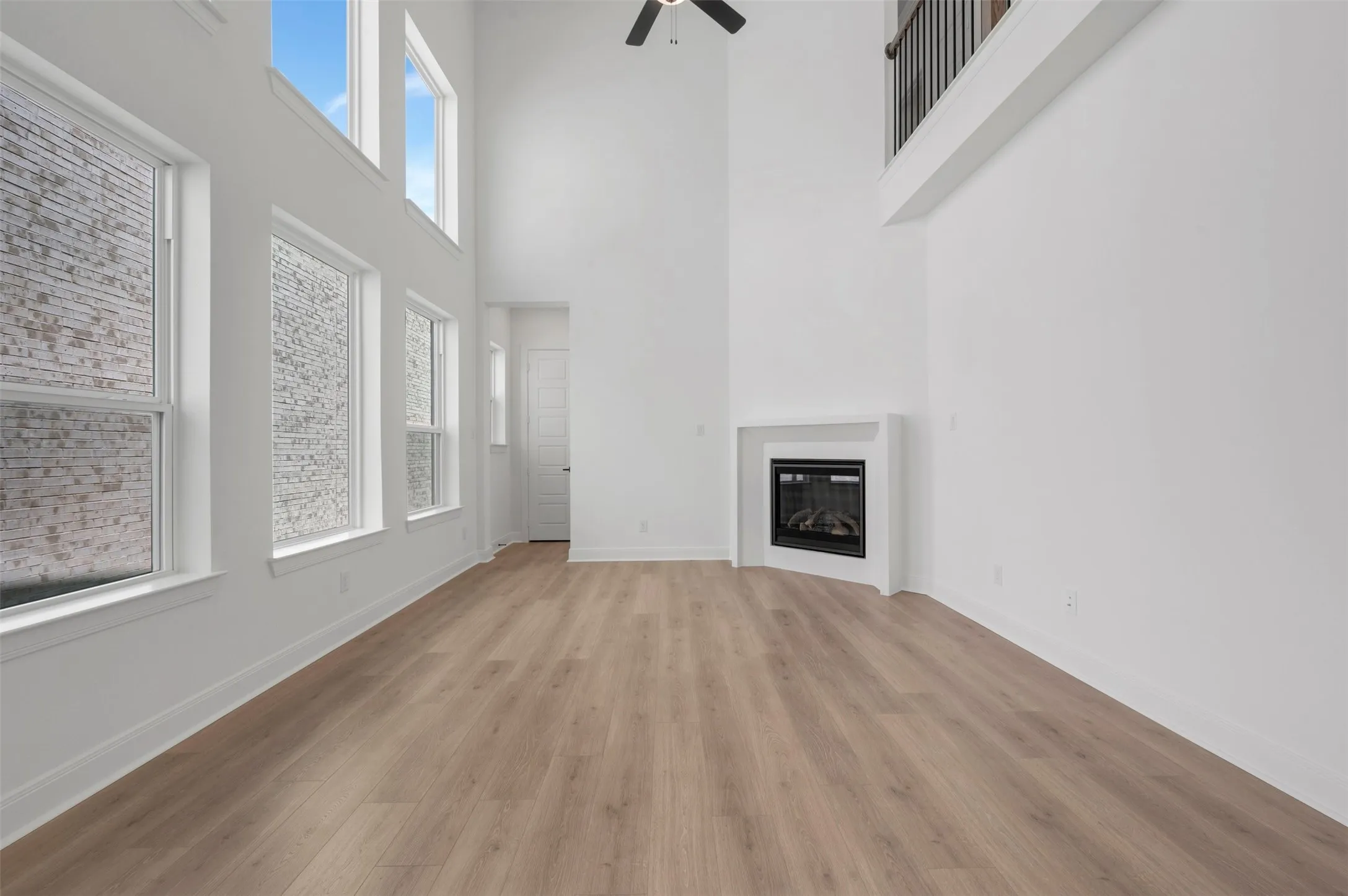 Soaring ceilings, abundant natural light from multiple windows, elegant wood-finish flooring, a contemporary fireplace, and an upper-level balcony with metal balusters