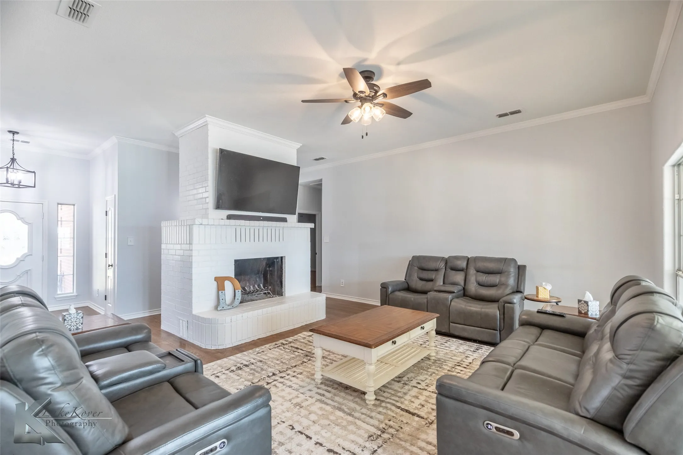 Living area featuring a ceiling fan, a brick fireplace, wood finished floors, crown molding, and a chandelier