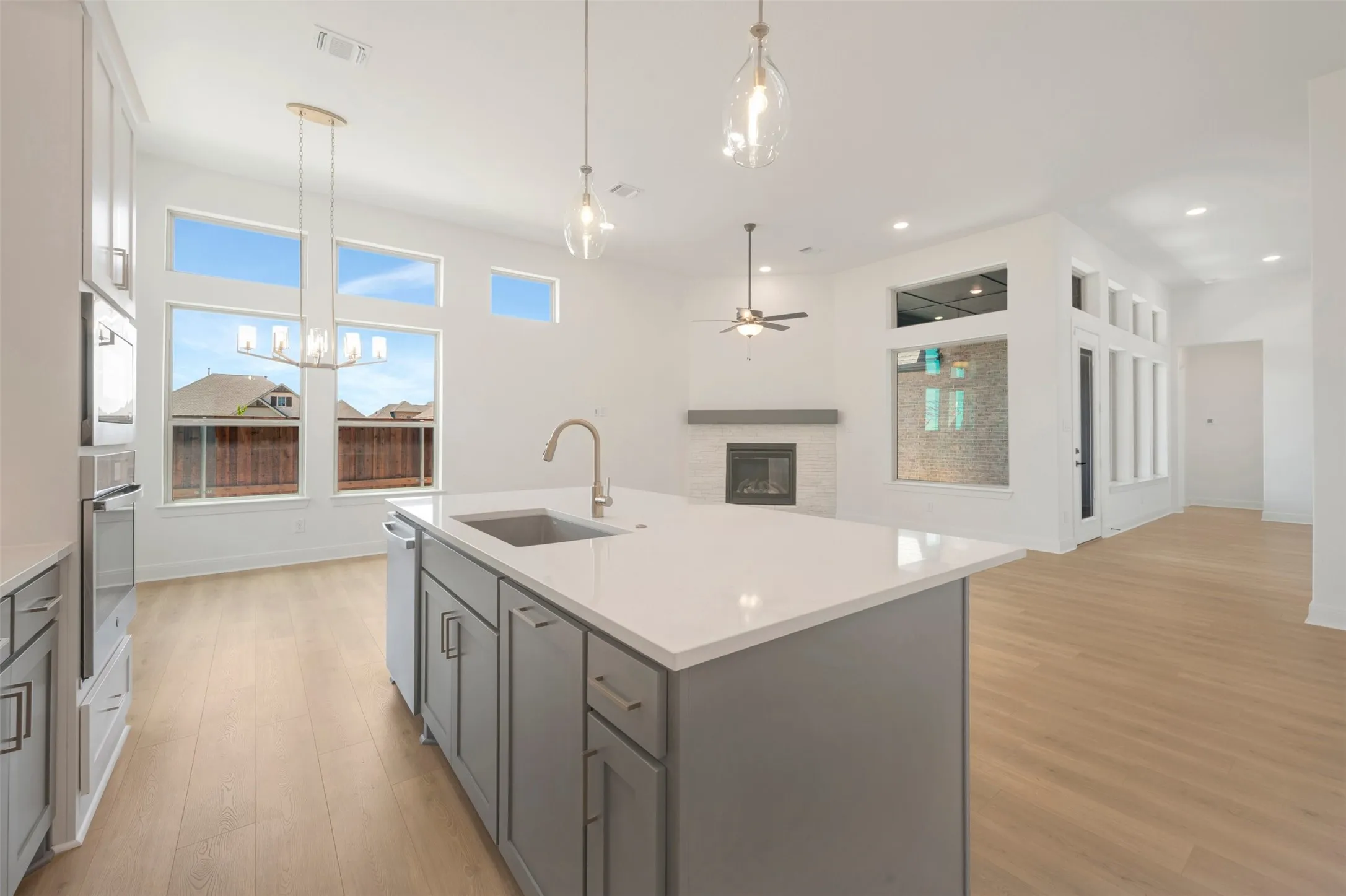 Open-concept kitchen featuring a spacious island with a sink, light wood-finish flooring, a contemporary chandelier, built-in oven, and a fireplace with a mantel