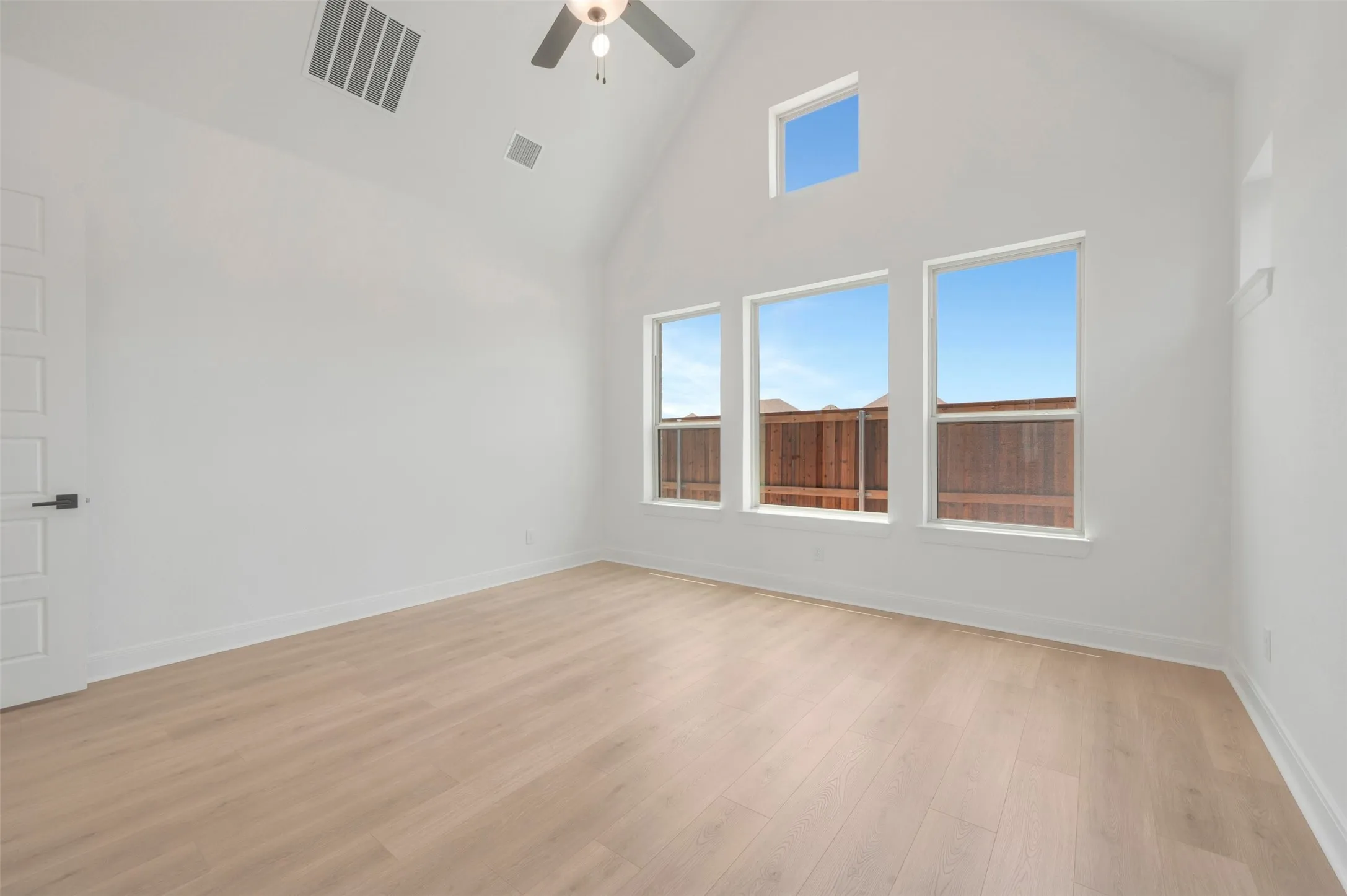 Vaulted ceiling with a ceiling fan, light wood-finish flooring, three large windows, and an additional high window