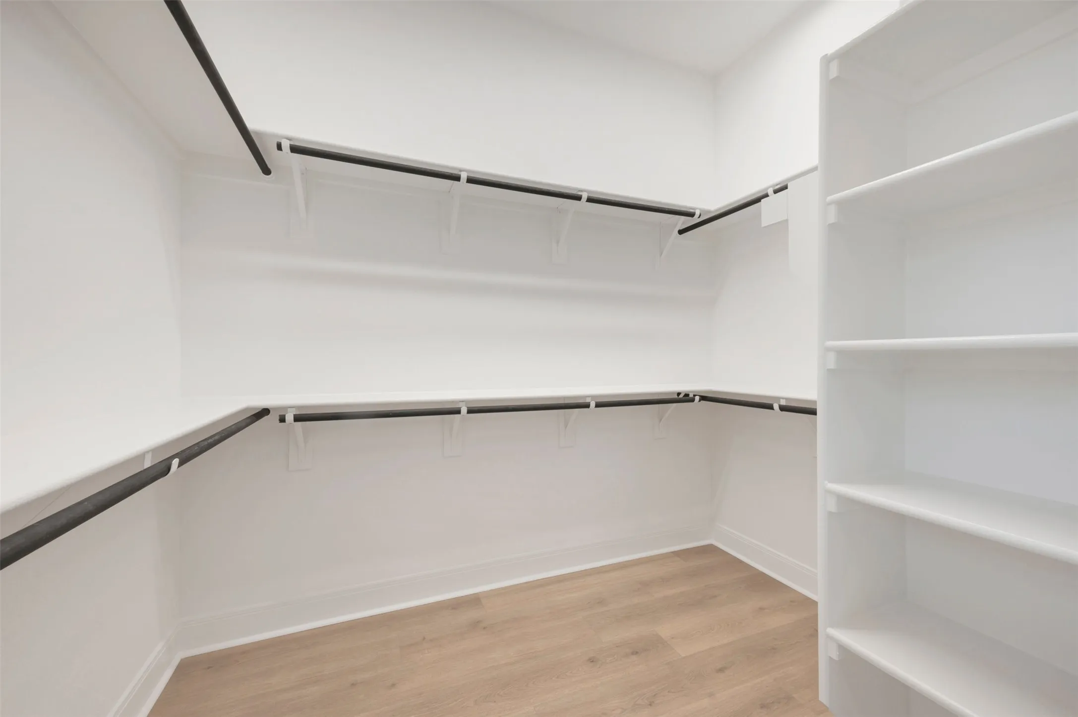 Spacious walk-in closet featuring light wood-finish flooring, white walls, built-in shelving, and multiple clothing rods