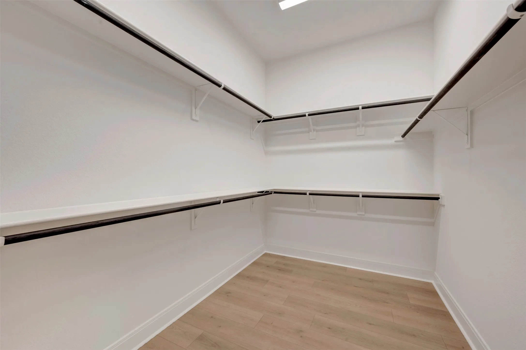Spacious walk-in closet featuring light wood-finish flooring, white walls, multiple storage shelves, and black hanging rods