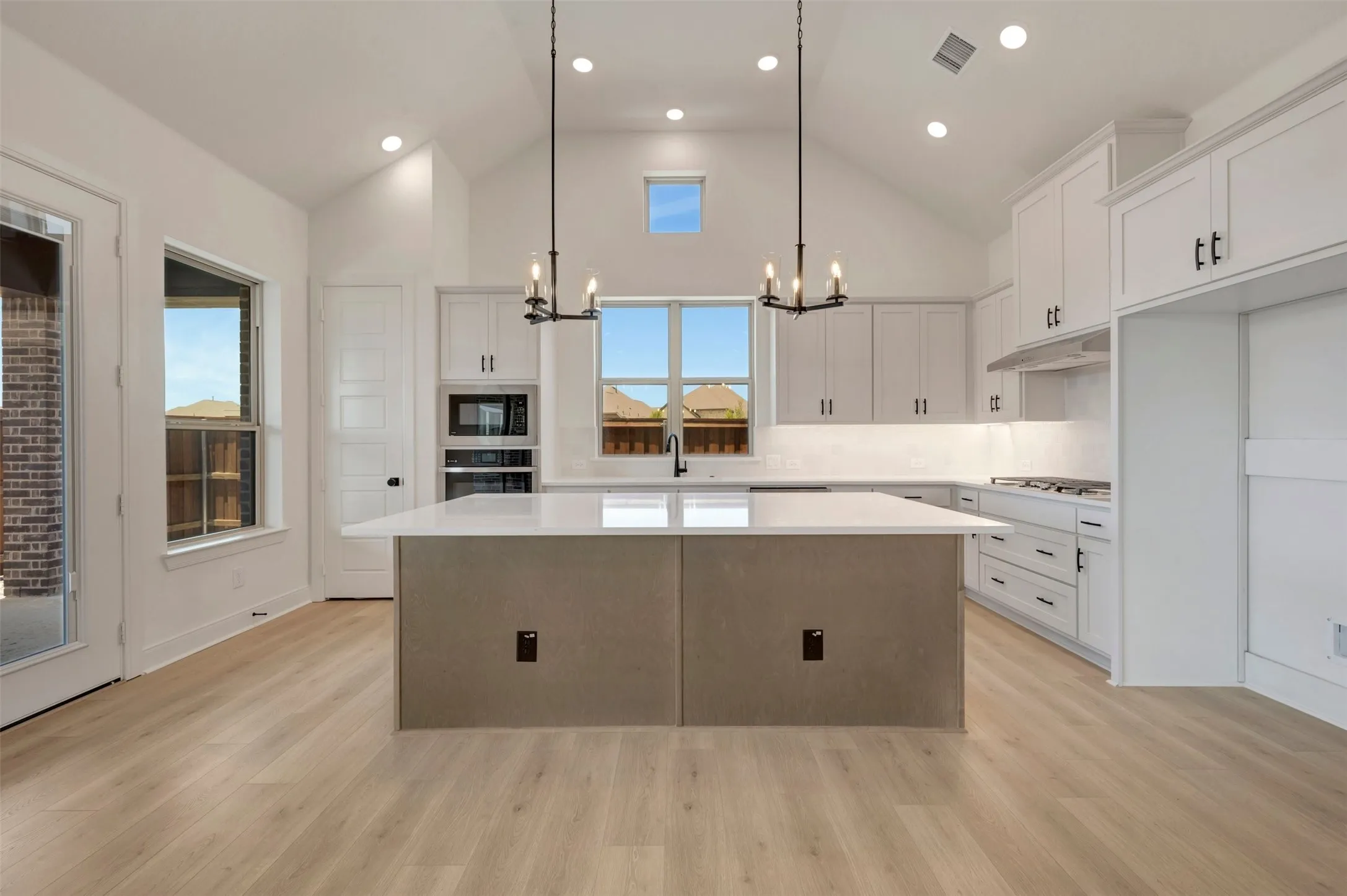 Spacious kitchen featuring a large central island with a light-toned countertop, ample white cabinetry with dark hardware, built-in double ovens, and a gas cooktop
