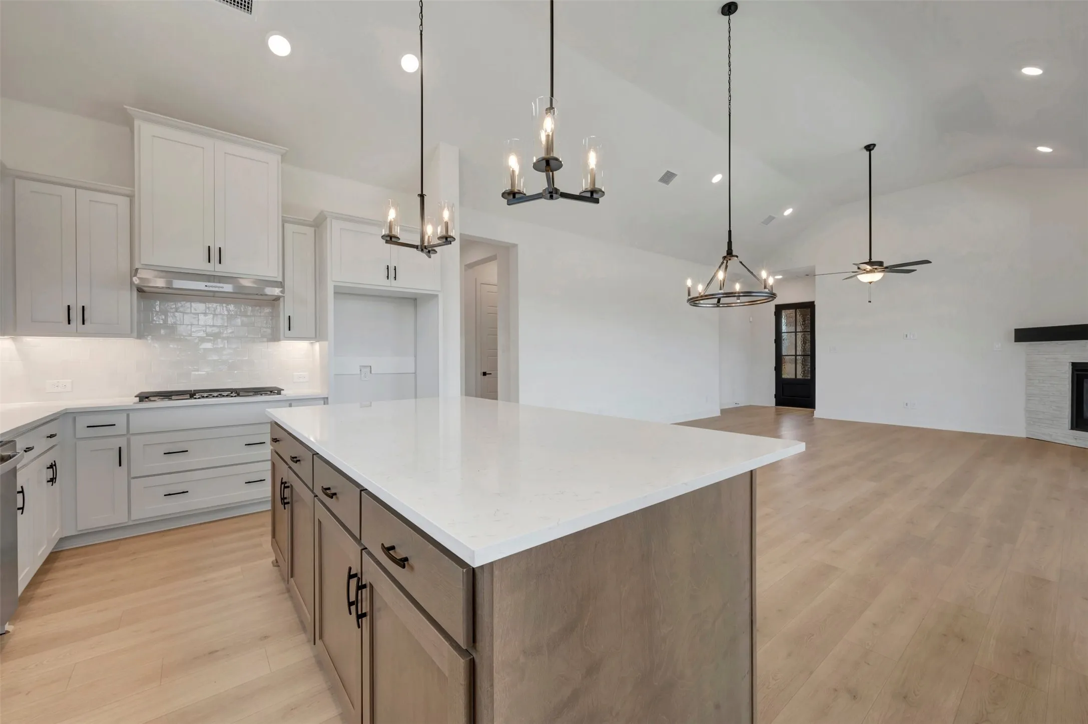 Open-concept living space featuring a spacious kitchen island with a light-toned countertop, white cabinetry with dark hardware, stainless steel appliances including a range hood, and wood-finish flooring