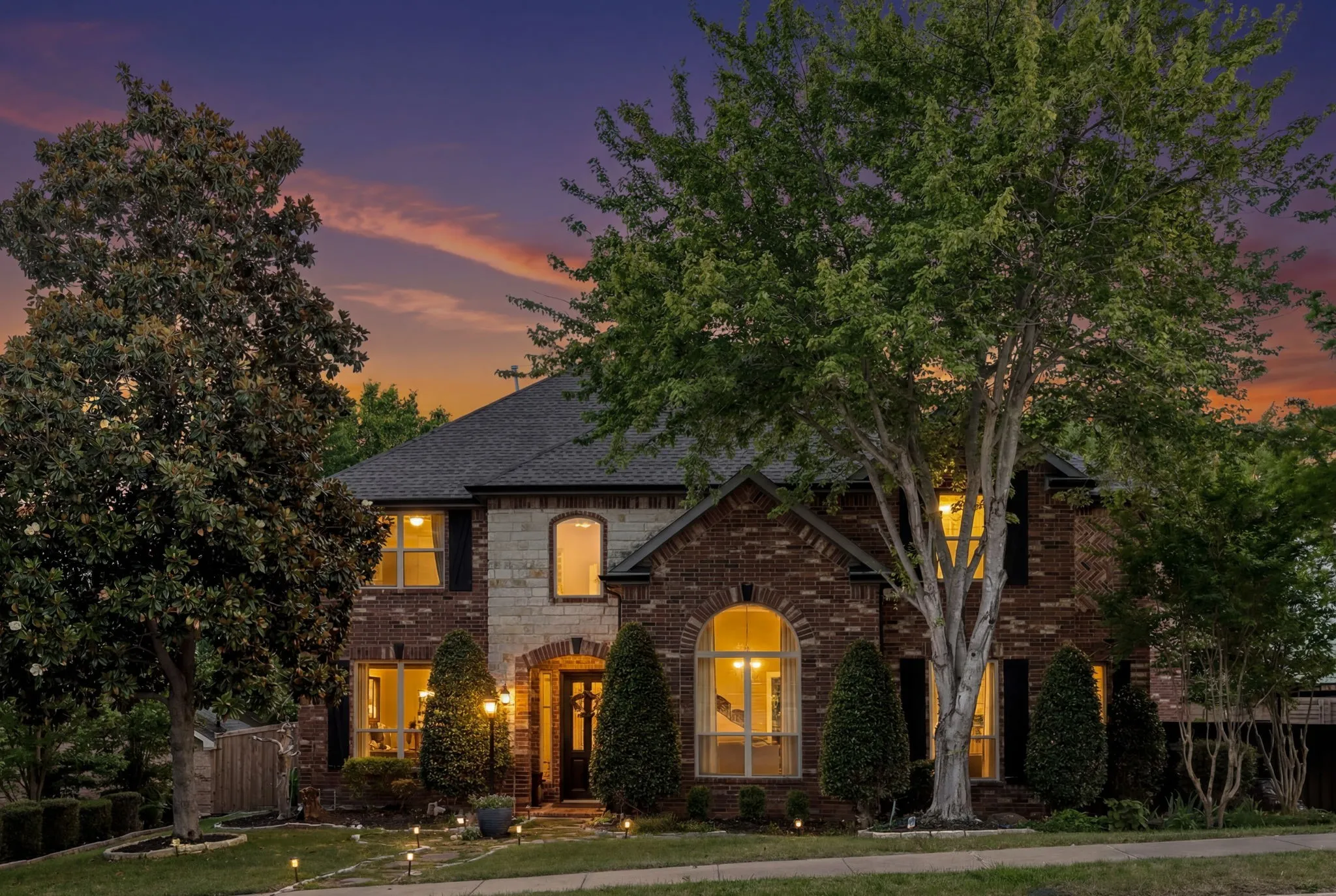 Brick and stone facade residence featuring a dark shingle roof and multiple arched windows