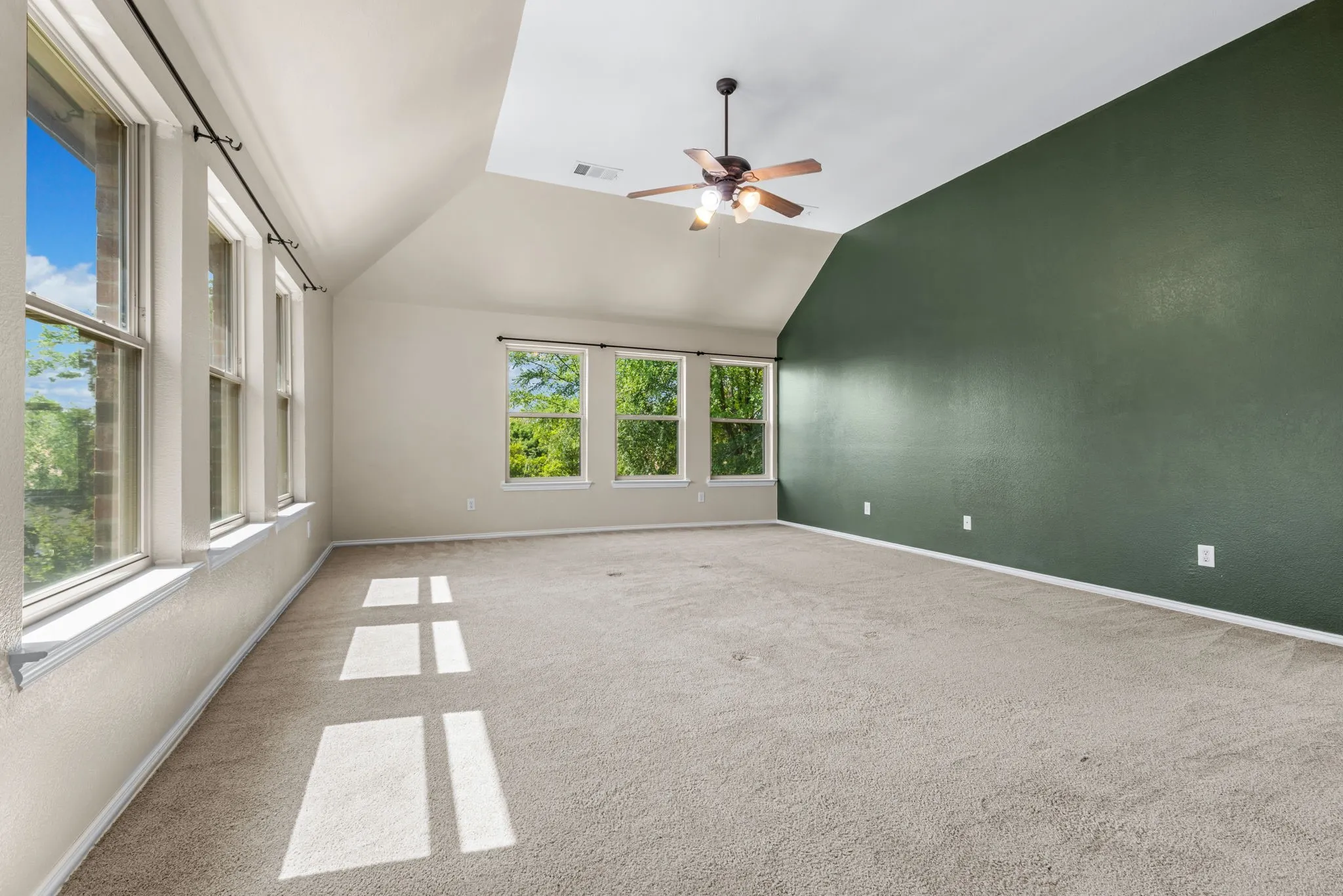 Spacious room featuring vaulted ceilings, a ceiling fan, plush carpet flooring, a dark green accent wall, and multiple windows