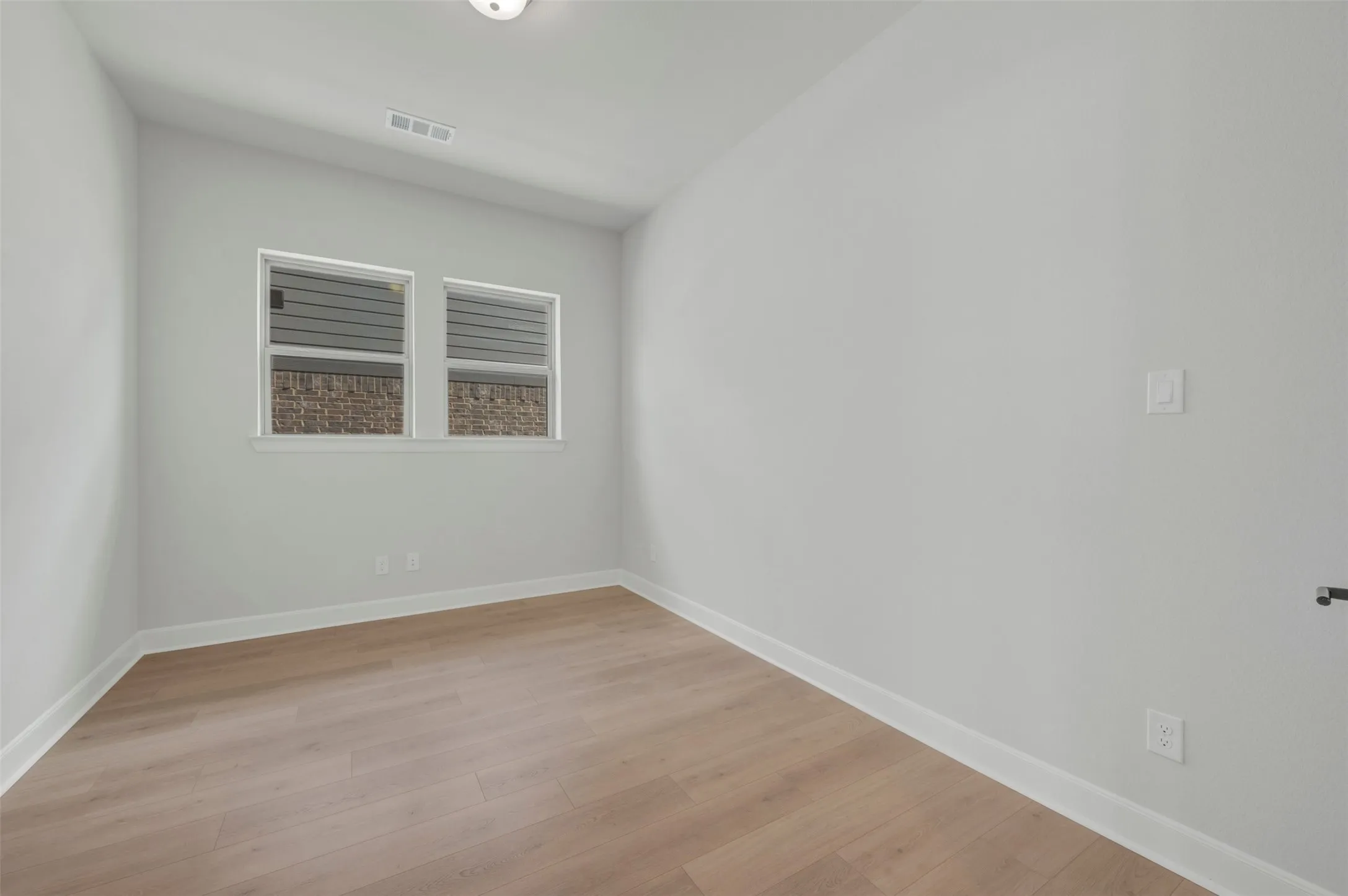 Bright room featuring wood-finish flooring, white baseboards, two windows with white trim, a flush-mount ceiling light, and a wall vent