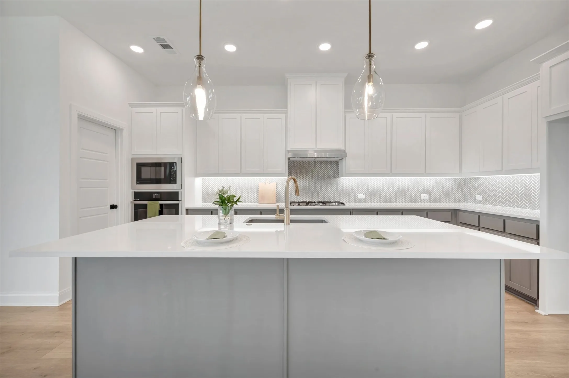 Spacious kitchen featuring a large central island with a white countertop, recessed lighting, pendant lighting, built-in stainless steel appliances, and a herringbone-patterned backsplash