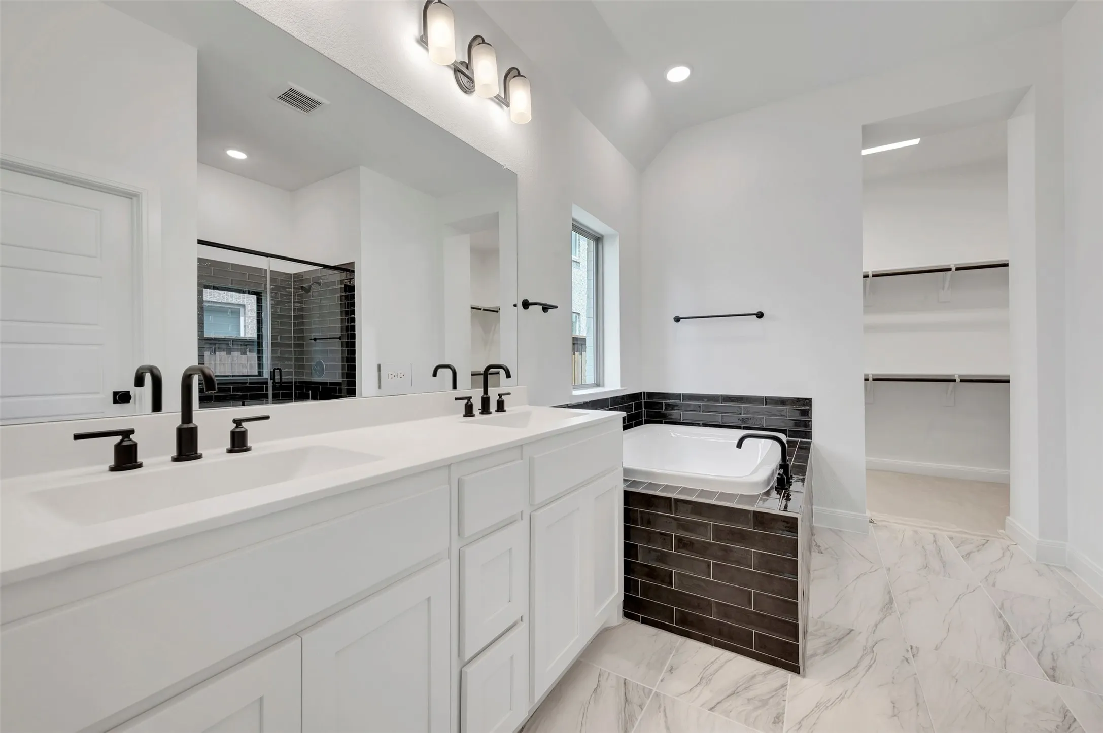 Spacious bathroom featuring a dual vanity with white cabinetry, matte black fixtures, a built-in soaking tub with dark subway tile surround, and large format marble-look floor tiles