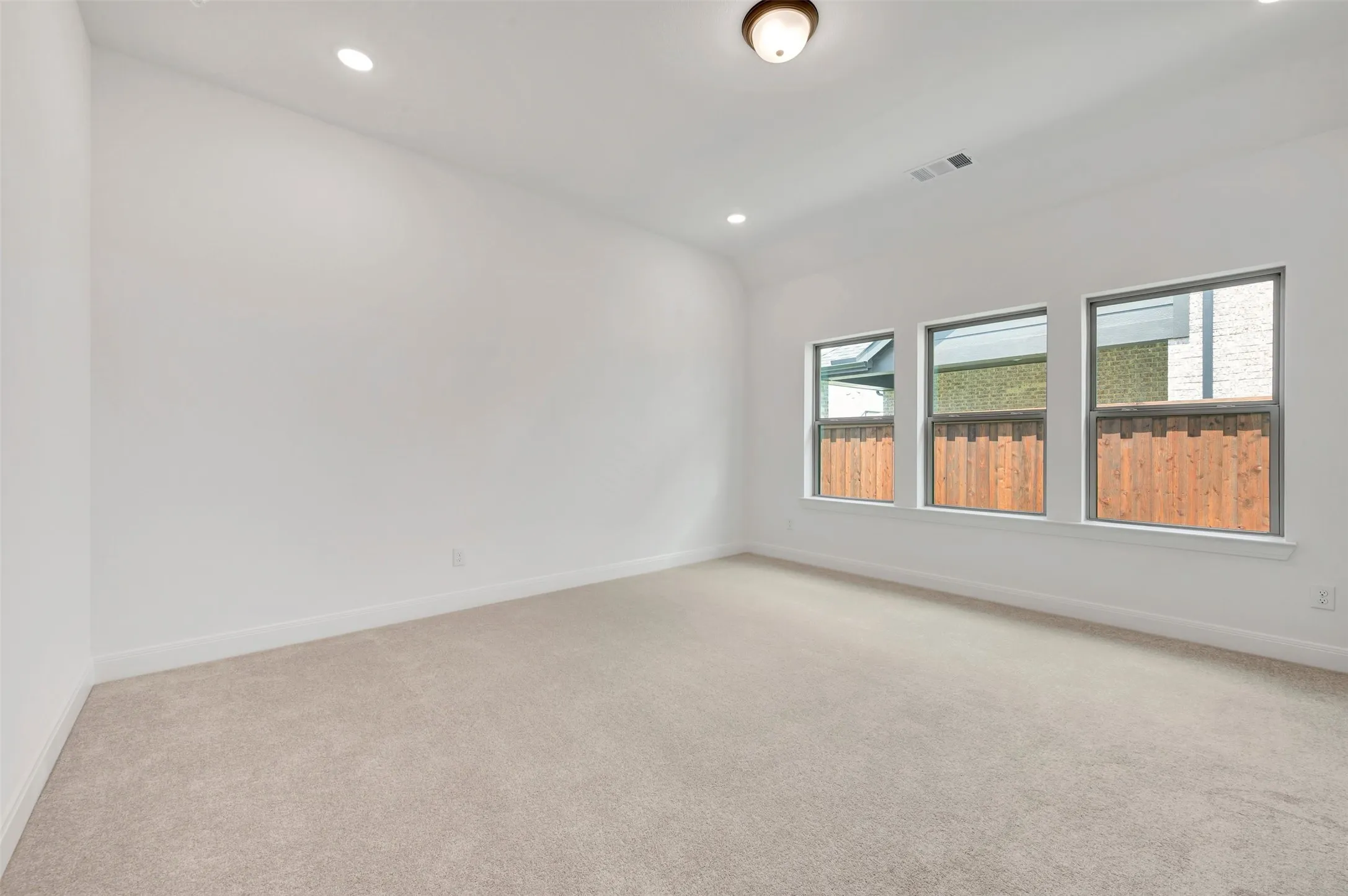 Spacious room featuring neutral carpeting, white walls, three windows with dark frames, recessed lighting, and a central ceiling light fixture