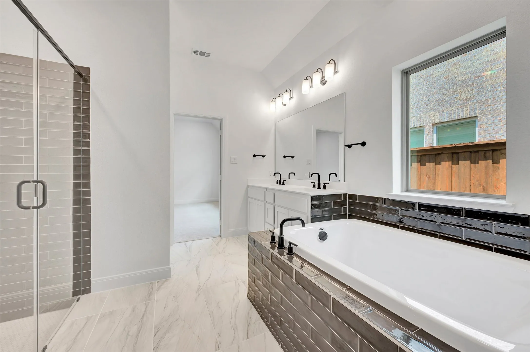 Contemporary bathroom featuring a spacious soaking tub with dark subway tile surround, a dual vanity with white cabinetry and dark fixtures, and a glass-enclosed shower with subway tile