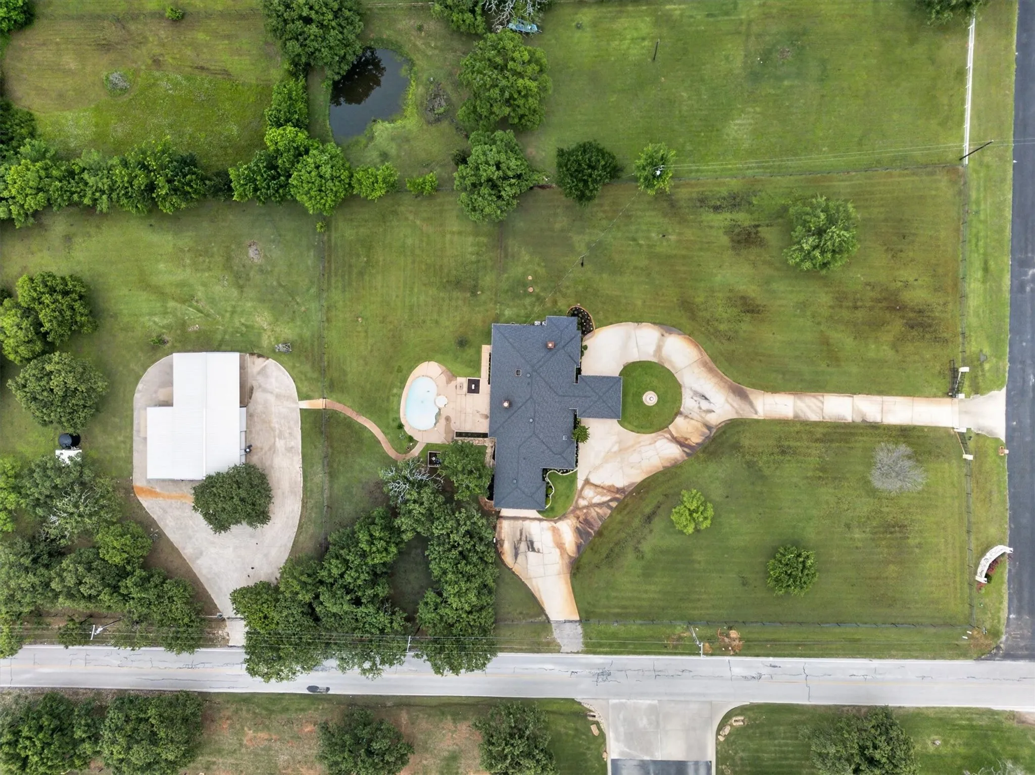Expansive property featuring a main residence with a dark shingle roof, a detached auxiliary building, a freeform swimming pool, and a circular driveway with a central landscaped island