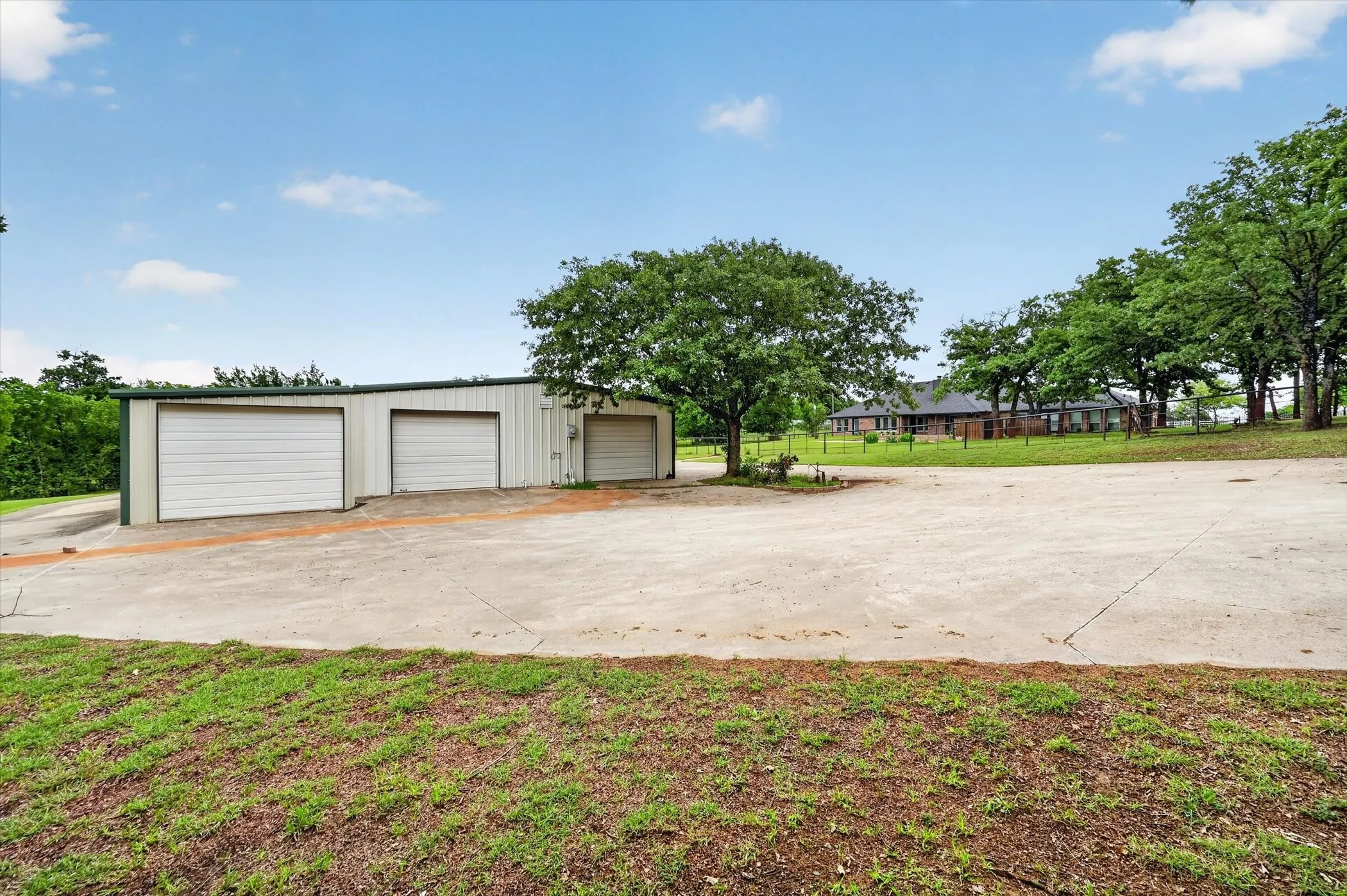 Multi-bay metal outbuilding featuring three roll-up garage doors, a concrete apron, and surrounding mature trees