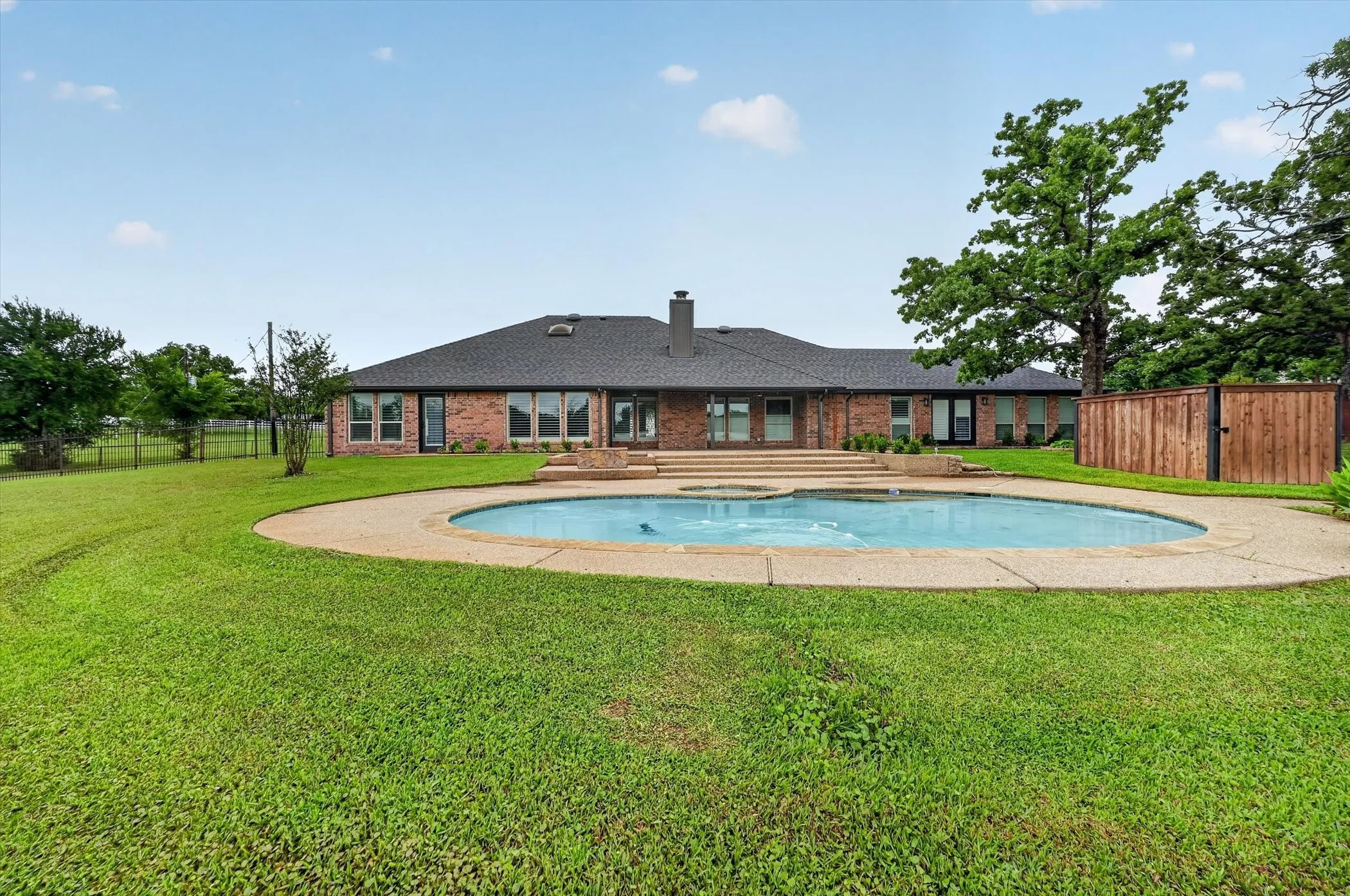 Expansive backyard featuring a large swimming pool with concrete surround, mature trees, and a brick exterior residence with multiple windows and a dark shingle roof