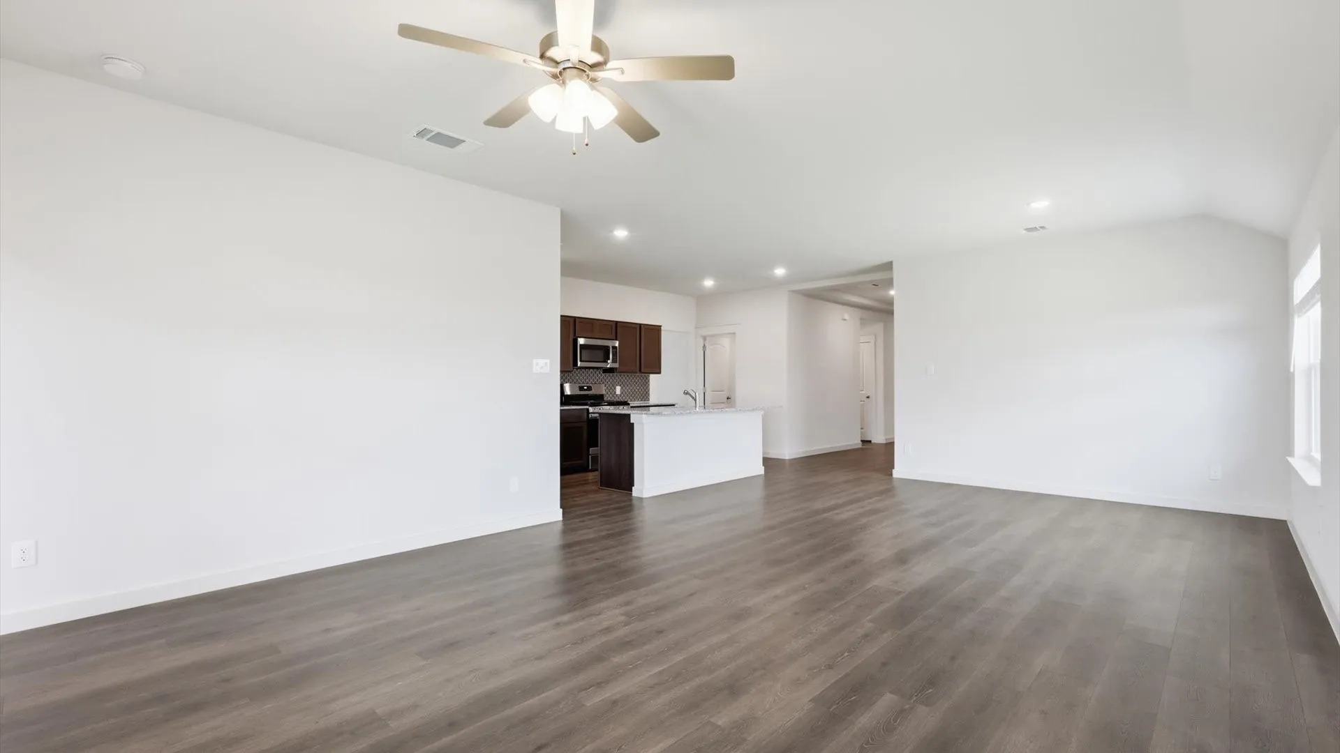 Spacious interior with wood-finish flooring, a contemporary ceiling fan, recessed lighting, and a kitchen featuring dark wood cabinetry, stainless steel appliances, and a white island with an undermount sink
