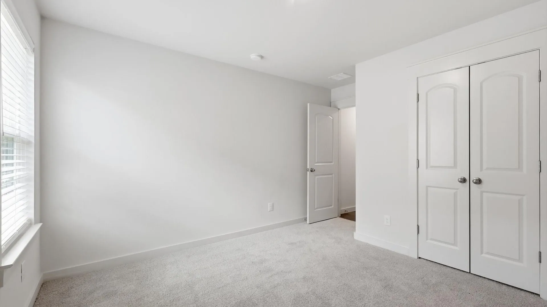Room featuring light gray carpet, white walls, a window with horizontal blinds, white baseboards, and two white arch-panel closet doors with round chrome hardware