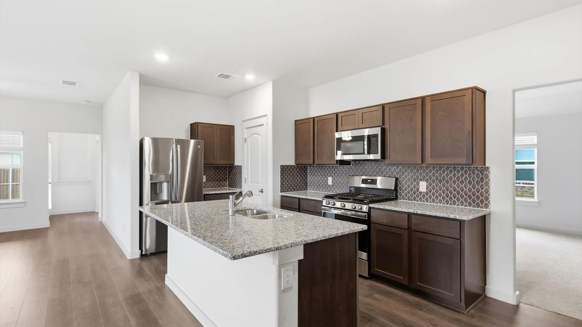 Modern kitchen featuring an island with a double basin sink, granite countertops, dark wood-finish cabinetry, stainless steel appliances, and a patterned tile backsplash