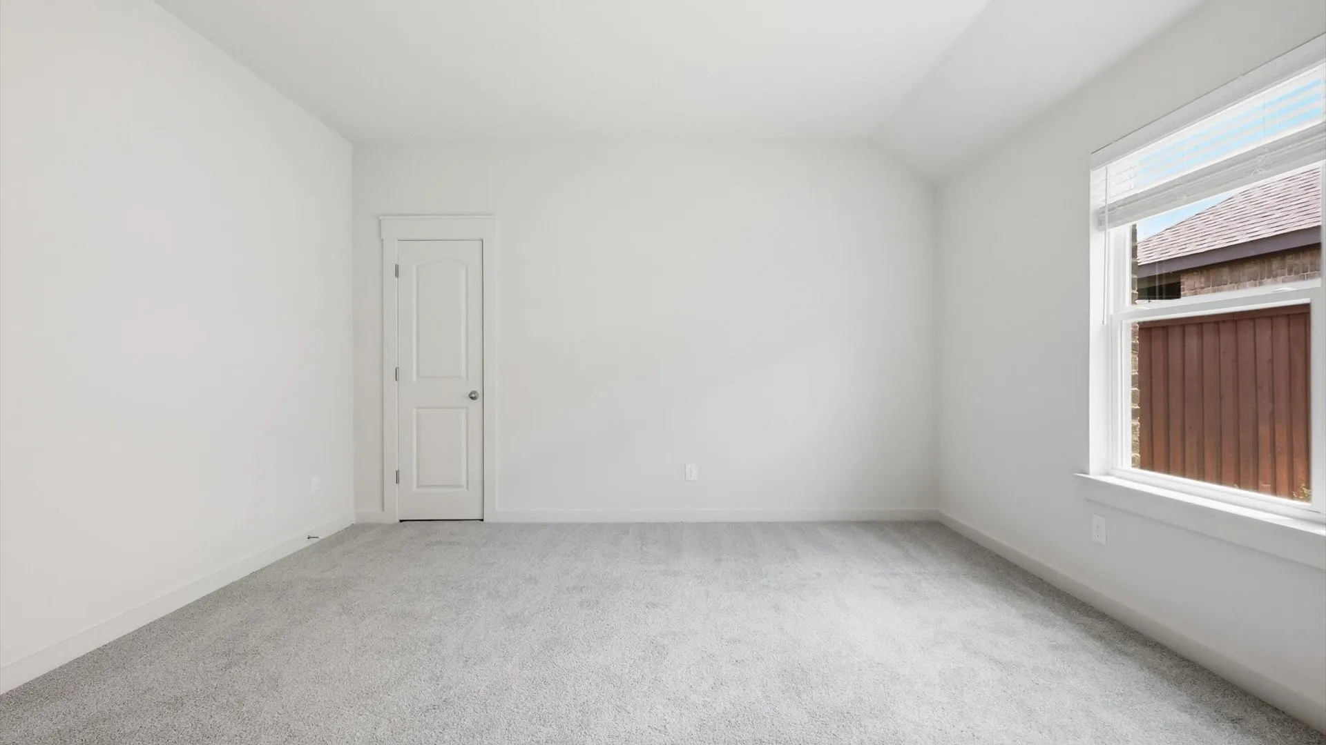 Spacious room featuring light gray carpet flooring, crisp white walls, a single window with blinds, and a white paneled door with a silver knob