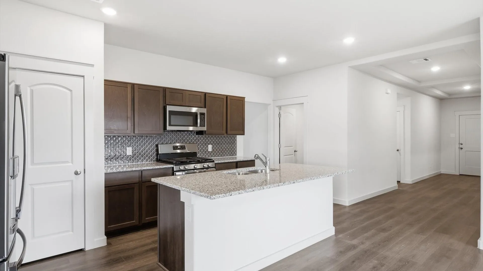 Open-concept kitchen featuring dark wood cabinetry, stainless steel appliances, a tile backsplash, and an island with a light-colored countertop and integrated sink