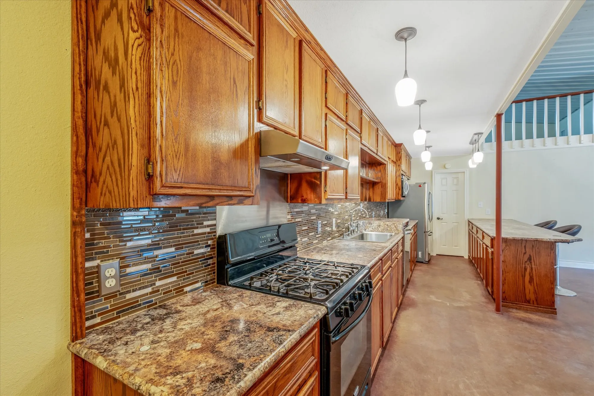 Kitchen with black gas stove, wood finish cabinets, decorative backsplash, decorative light fixtures, and light stone counters