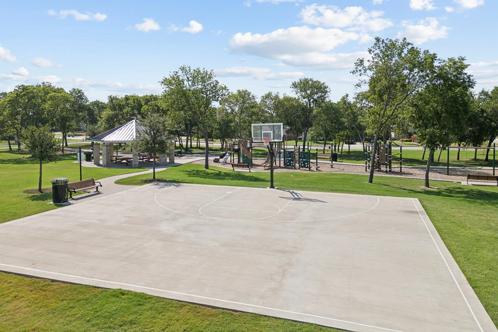 Basketball court within a park setting featuring a covered picnic area, playground equipment, and mature trees