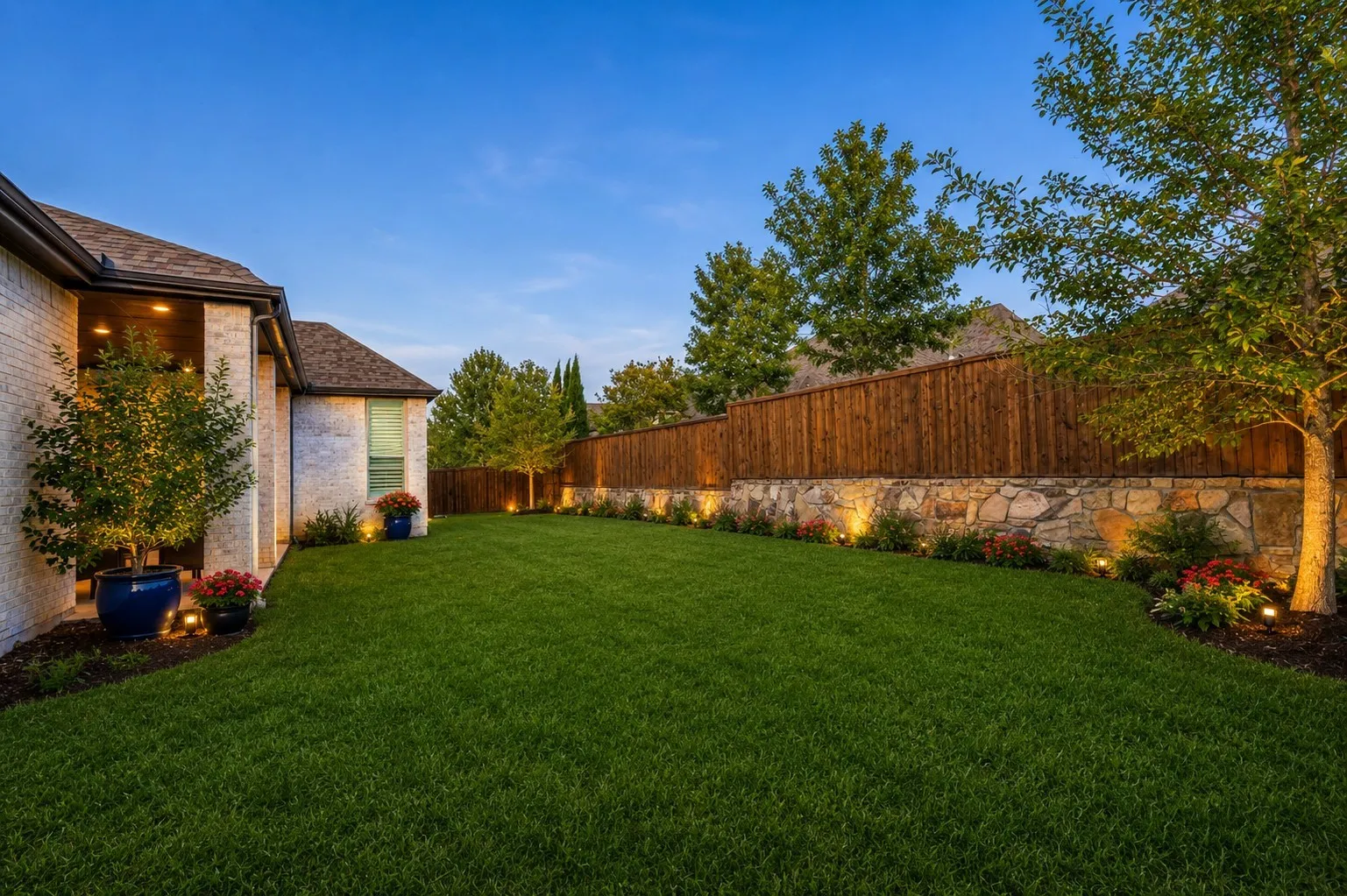 Spacious backyard featuring a manicured lawn, stone retaining wall, wood privacy fencing, and extensive landscape lighting. AI enhanced