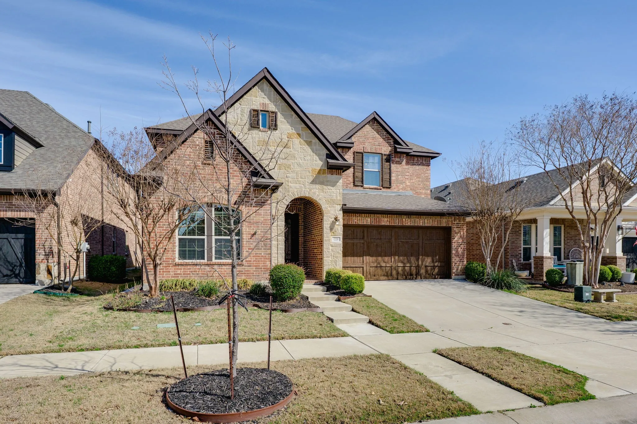 French country inspired facade featuring brick siding, concrete driveway, stone siding, a shingled roof, and a front lawn