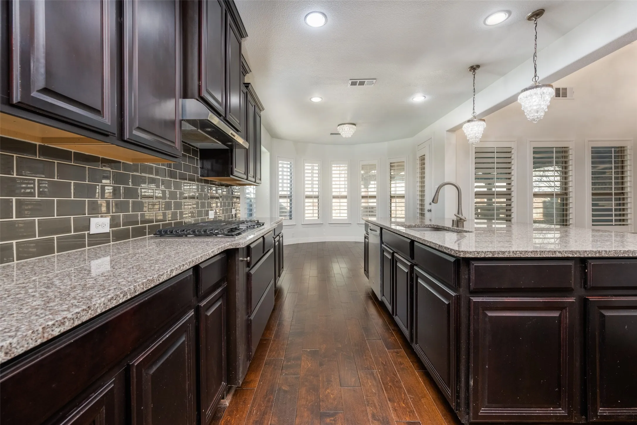 Kitchen with dark wood finish cabinetry, light stone countertops, dark wood-type flooring, tasteful backsplash, and a kitchen island with sink