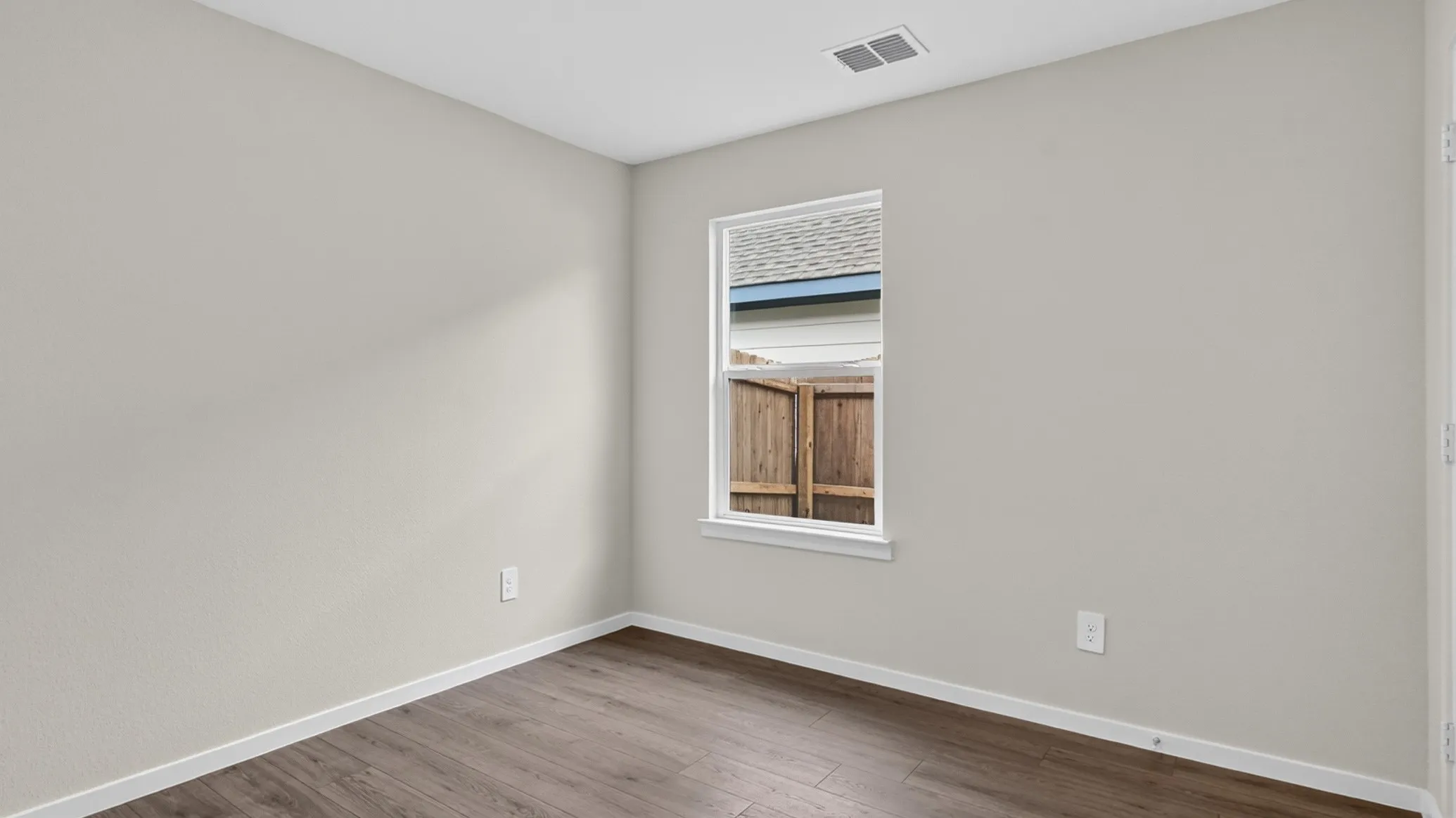 Neutral wall finishes and rich wood-finish flooring define this bright interior space, featuring a single window with white trim, crisp white baseboards, and a ceiling vent