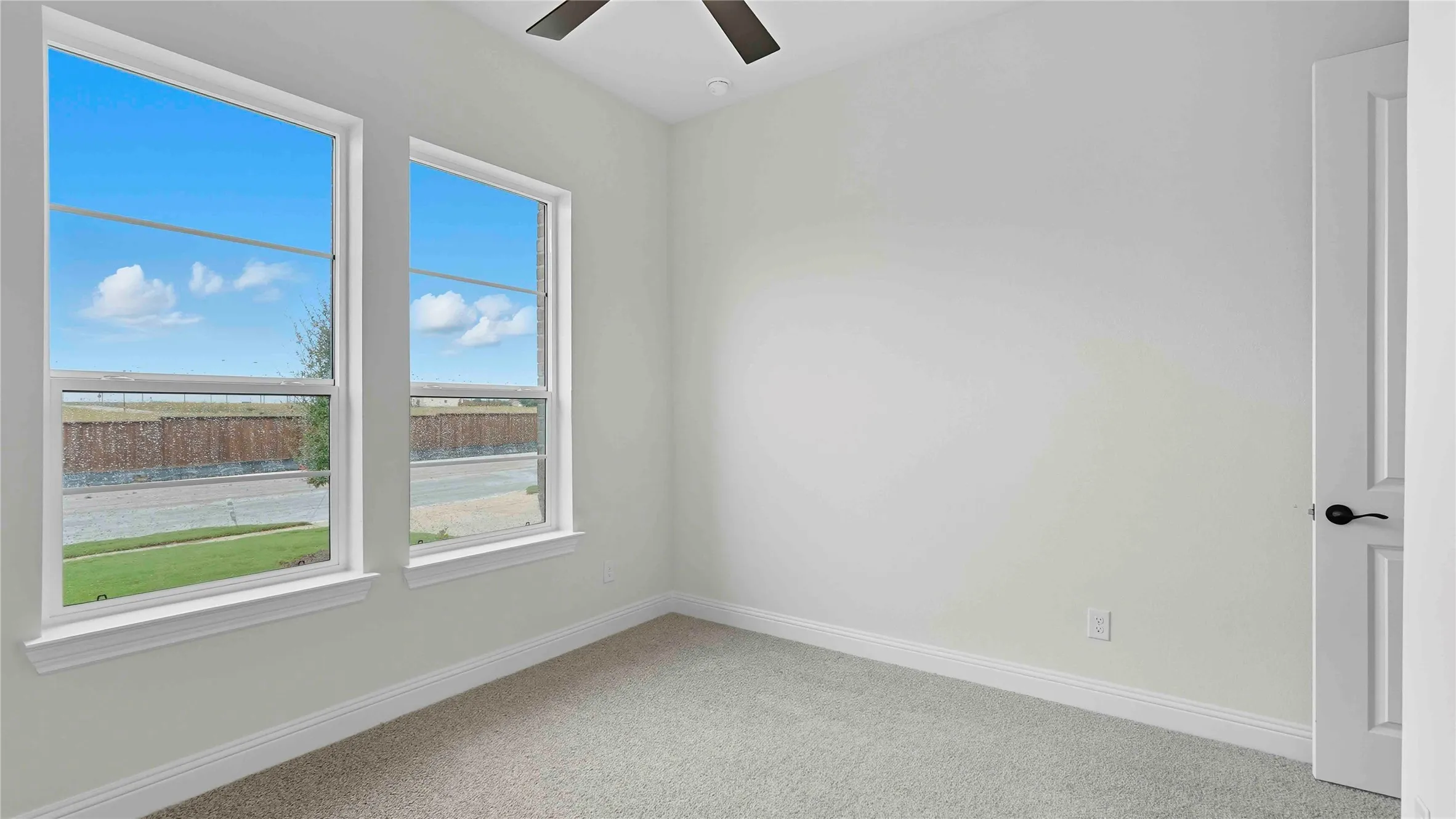 Bright room featuring two large windows, neutral-toned carpeting, white baseboards, and a ceiling fan with a dark finish