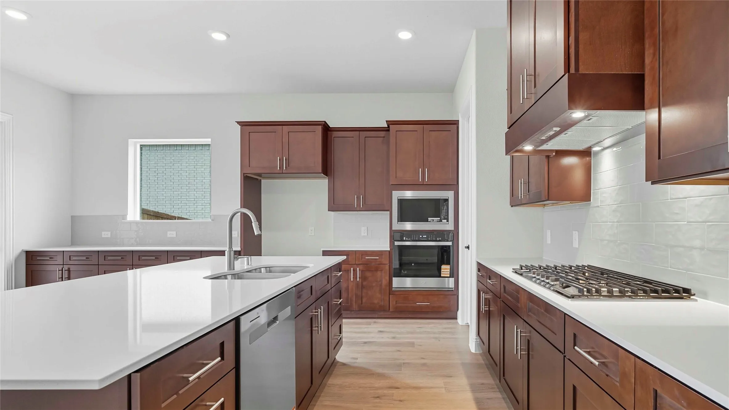 Kitchen featuring a large central island with an undermount sink and dishwasher, rich wood-finish cabinetry, light-toned countertops, and a white subway tile backsplash