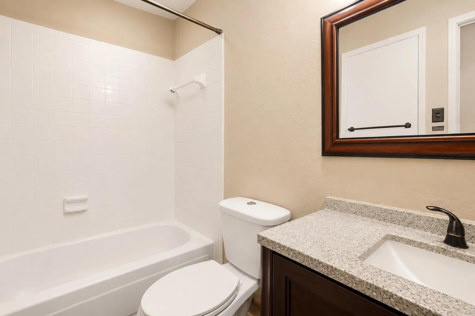 Bathroom featuring a bathtub with white tile surround, a toilet, a vanity with a light-colored speckled countertop, an undermount sink, and a dark bronze faucet