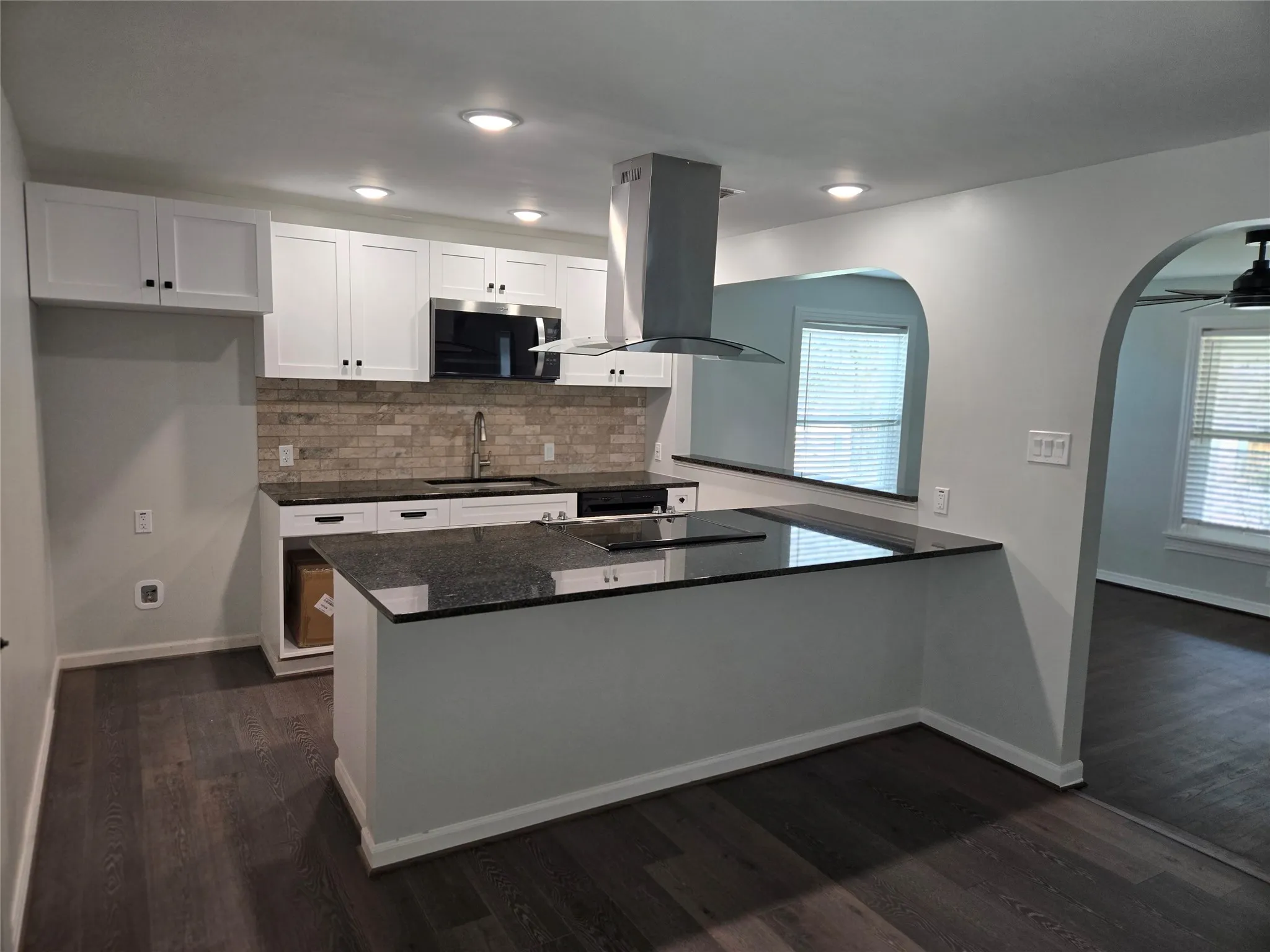 Kitchen featuring island range hood, white cabinets, a peninsula, decorative backsplash, and dark stone countertops