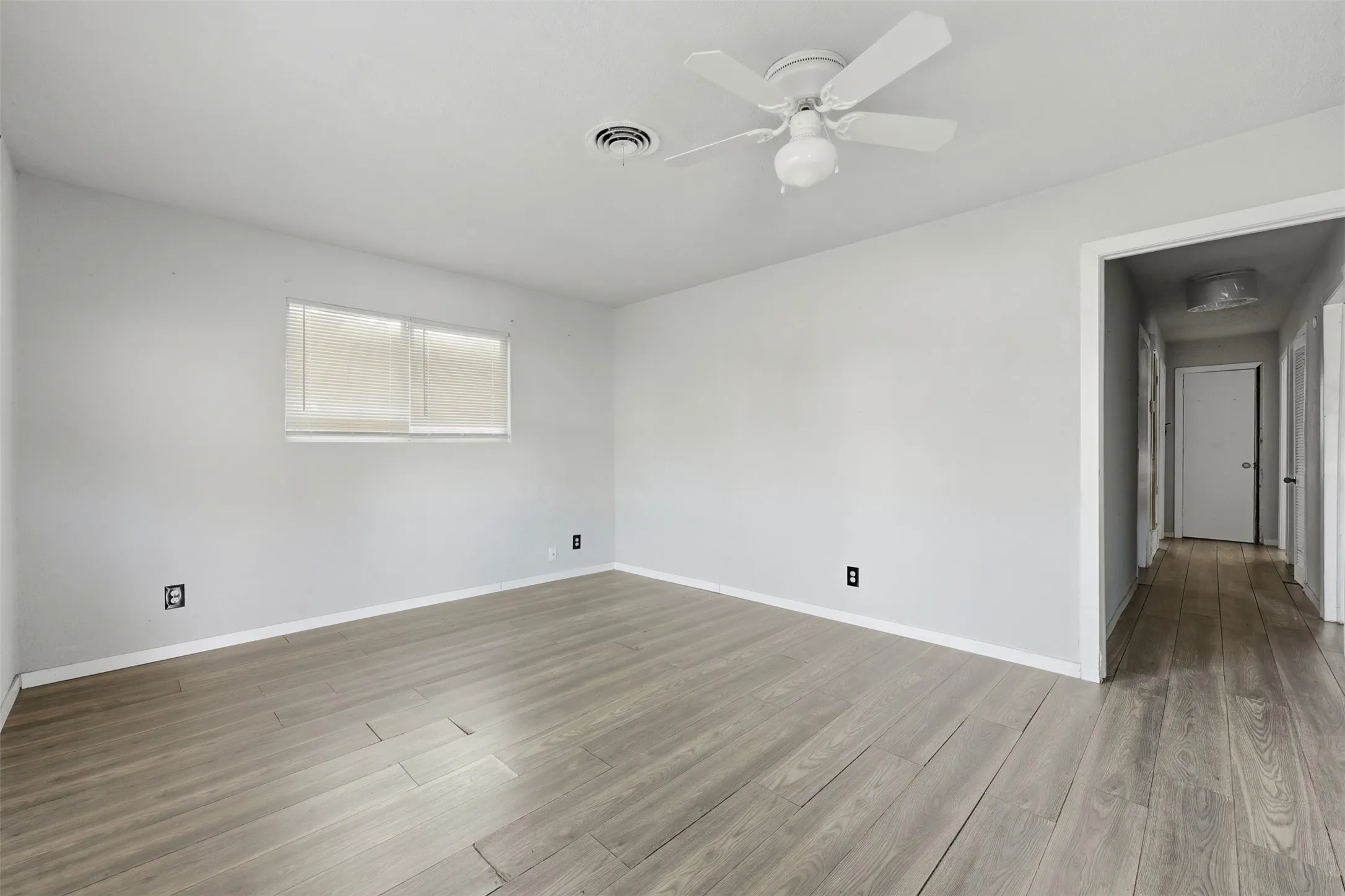 Light-toned wood-finish flooring, white walls, white ceiling fan, and a single window with blinds
