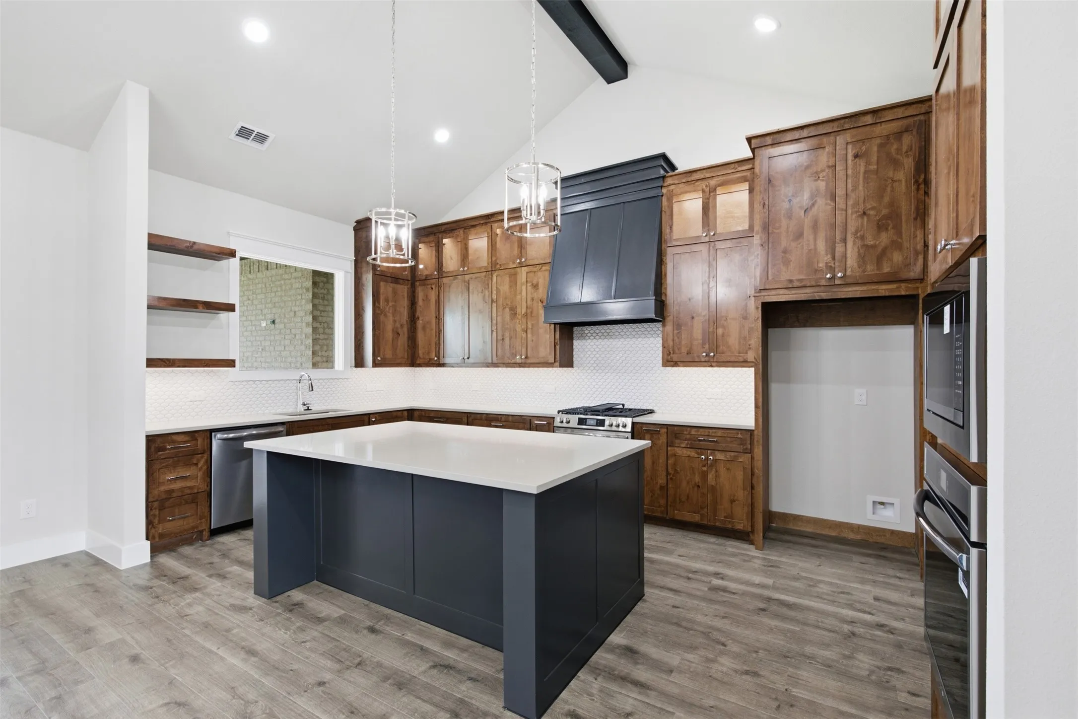 Kitchen featuring a large central island with a light-toned countertop, wood-finish flooring, dark wood cabinetry, a white herringbone tile backsplash, and stainless steel appliances