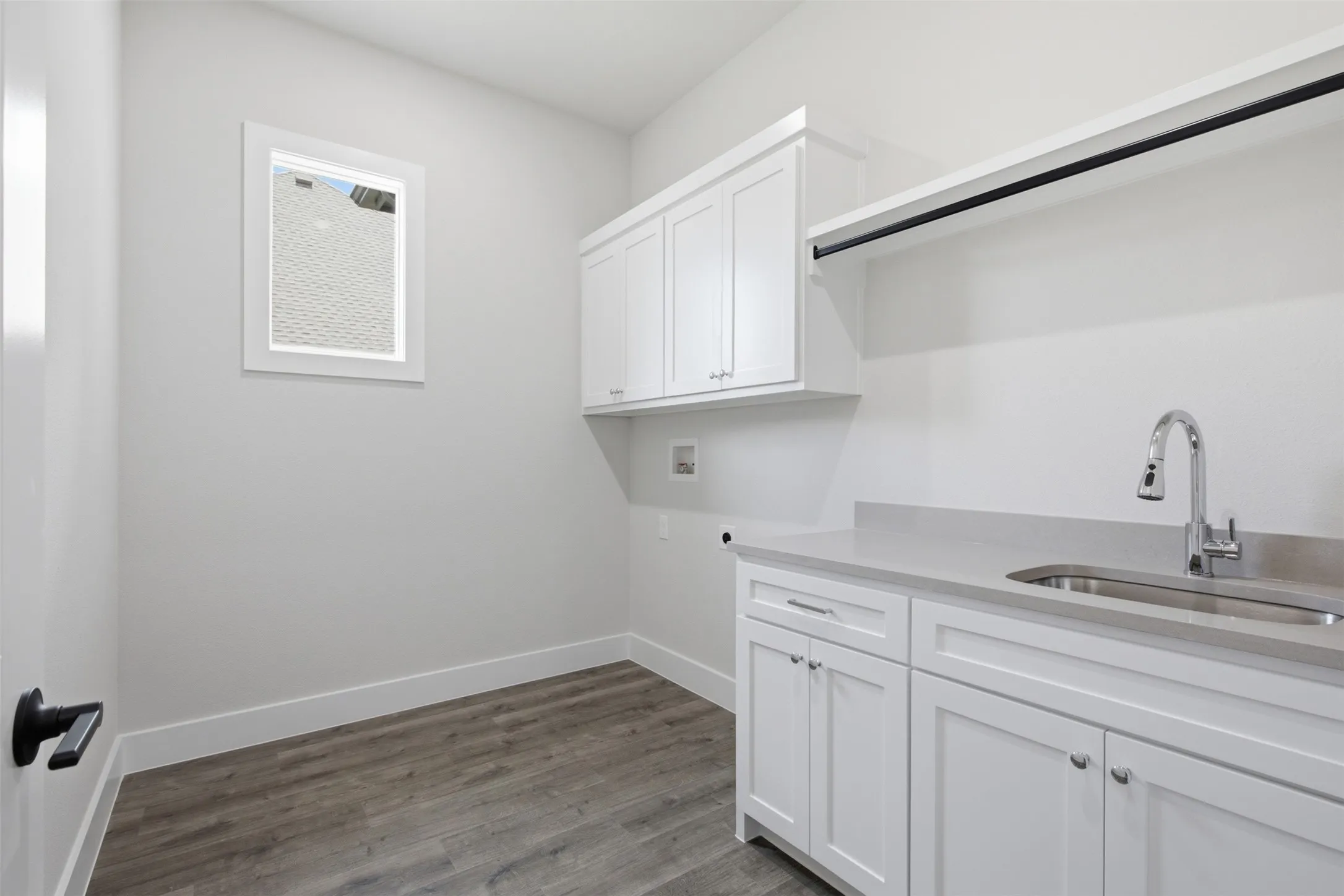 Dedicated laundry area featuring white shaker-style cabinetry, a stainless steel sink with a chrome gooseneck faucet, light-toned countertop, and wood-finish flooring