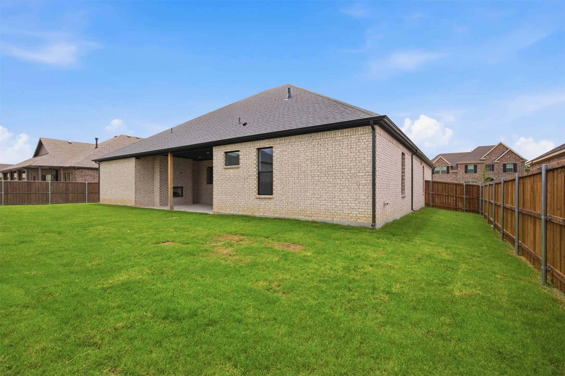 Expansive backyard featuring a lush green lawn, full privacy fencing, light-colored brick exterior, a covered patio area, and a dark shingle roof