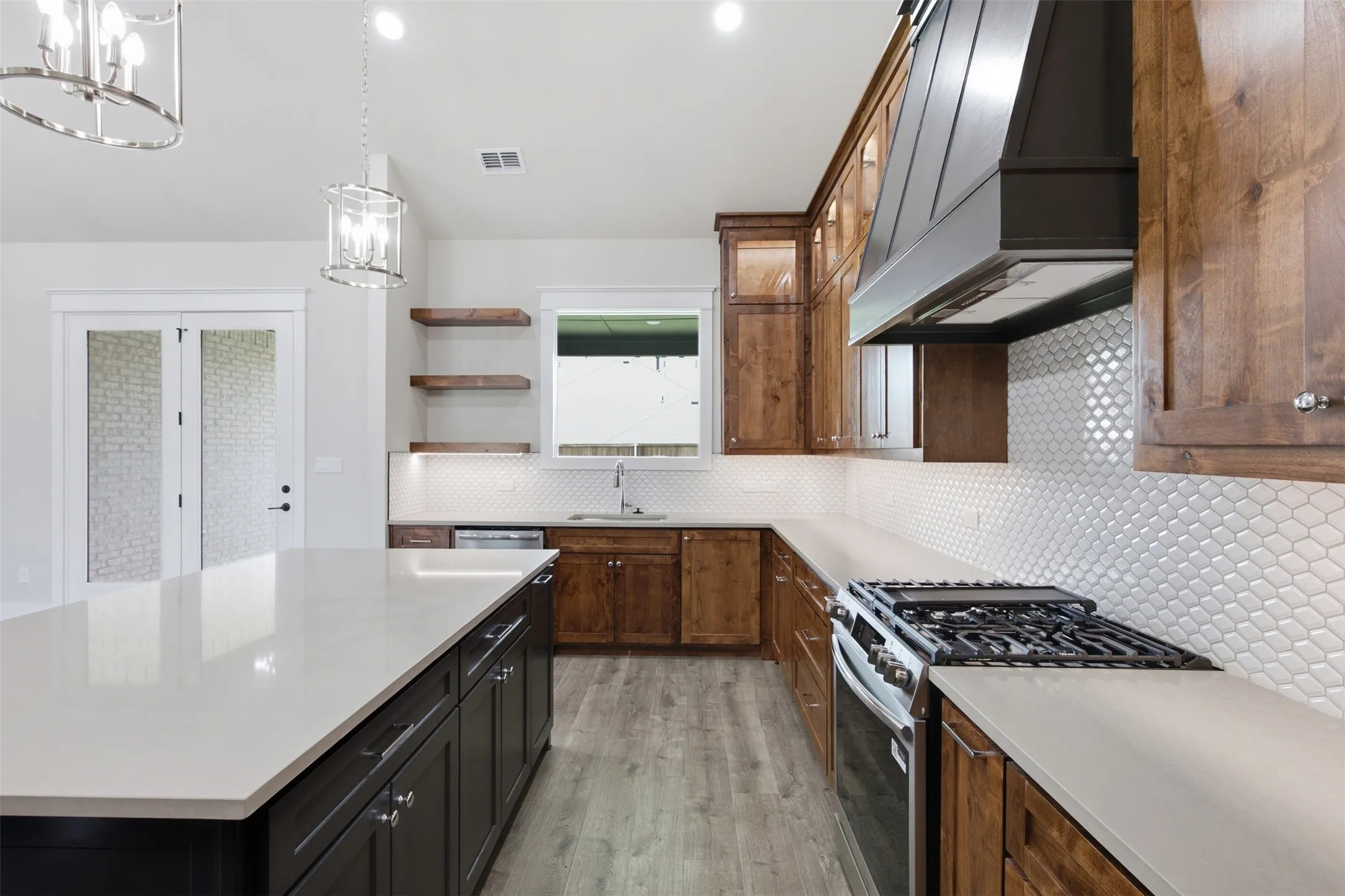 Gourmet kitchen featuring an expansive island with a light-toned countertop, a gas range with a substantial range hood, white scale-patterned backsplash, wood-finish flooring, and a combination of dark and natural wood cabinetry