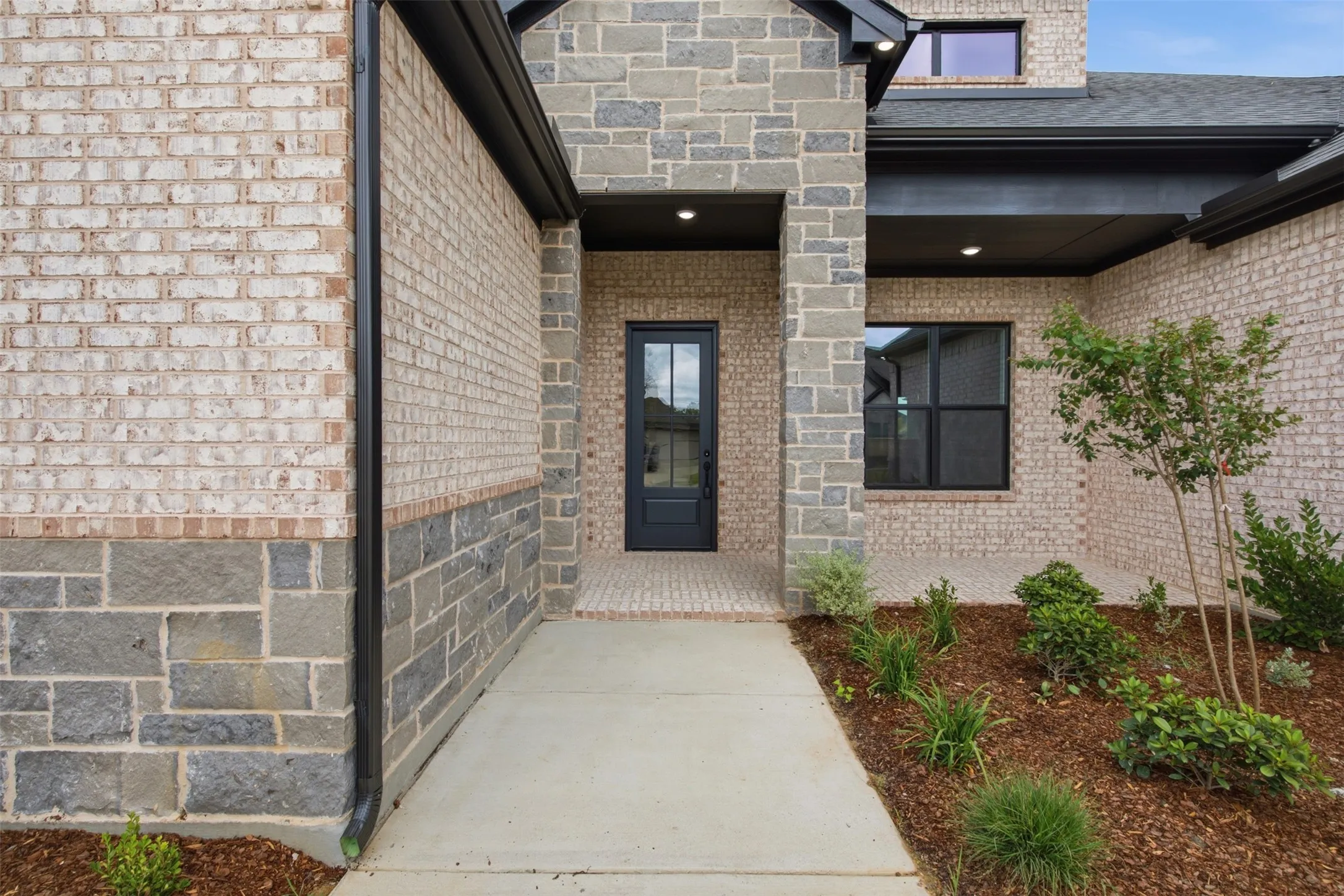 Entryway featuring a dark paneled door with glass inserts, brick and stone exterior, recessed lighting, and a landscaped garden bed