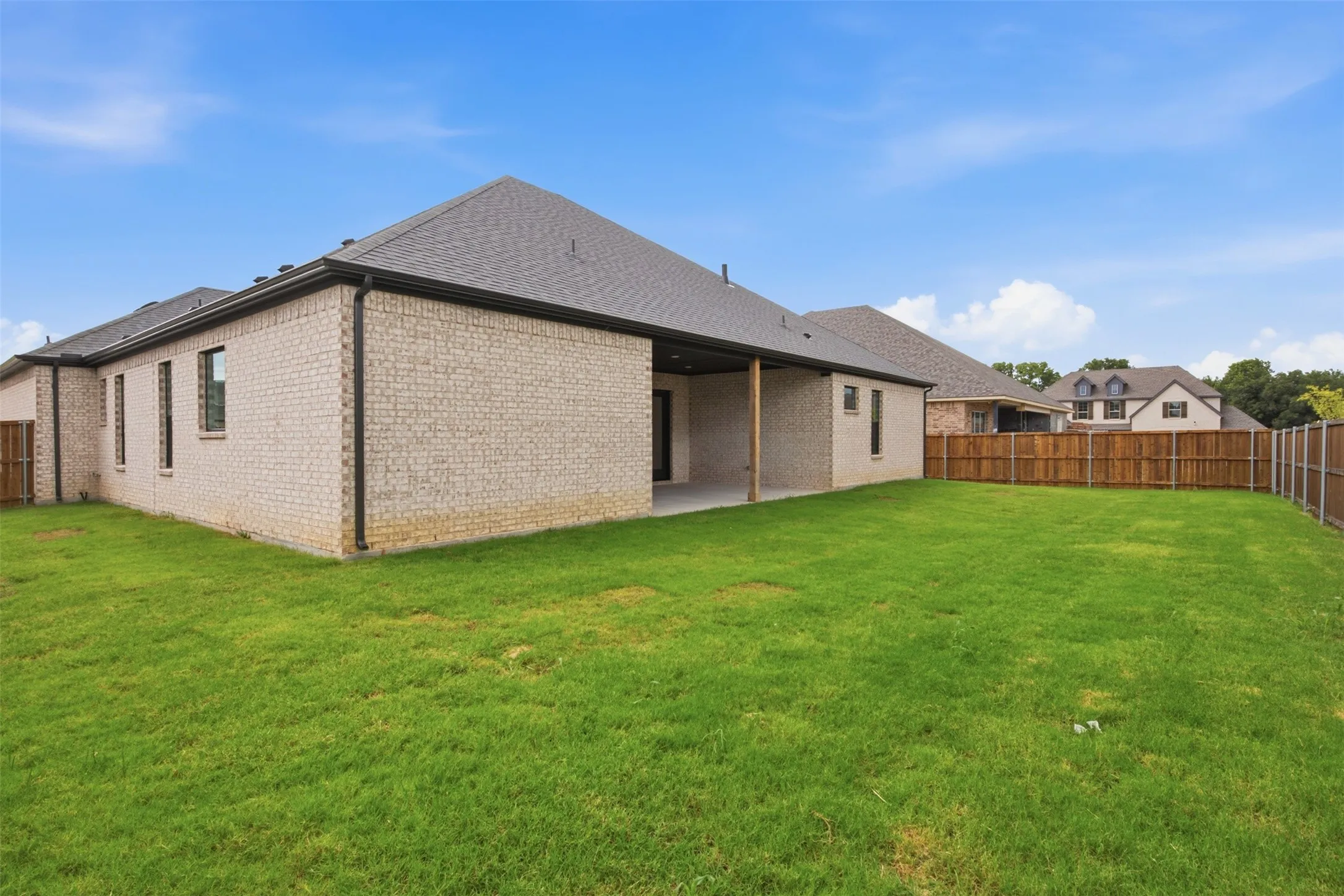 Expansive backyard featuring a green lawn, wood privacy fencing, a covered concrete patio, and light-colored brick exterior